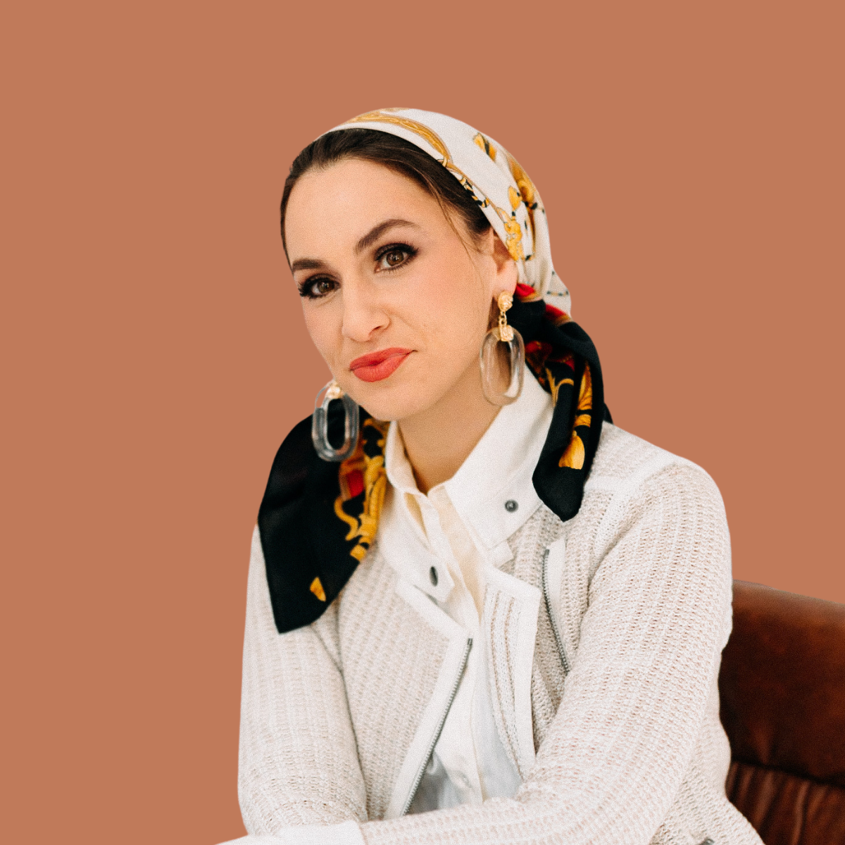 A woman with dark hair wearing a patterned headscarf, dangling earrings, and a white knit sweater, sitting against a plain brown background.