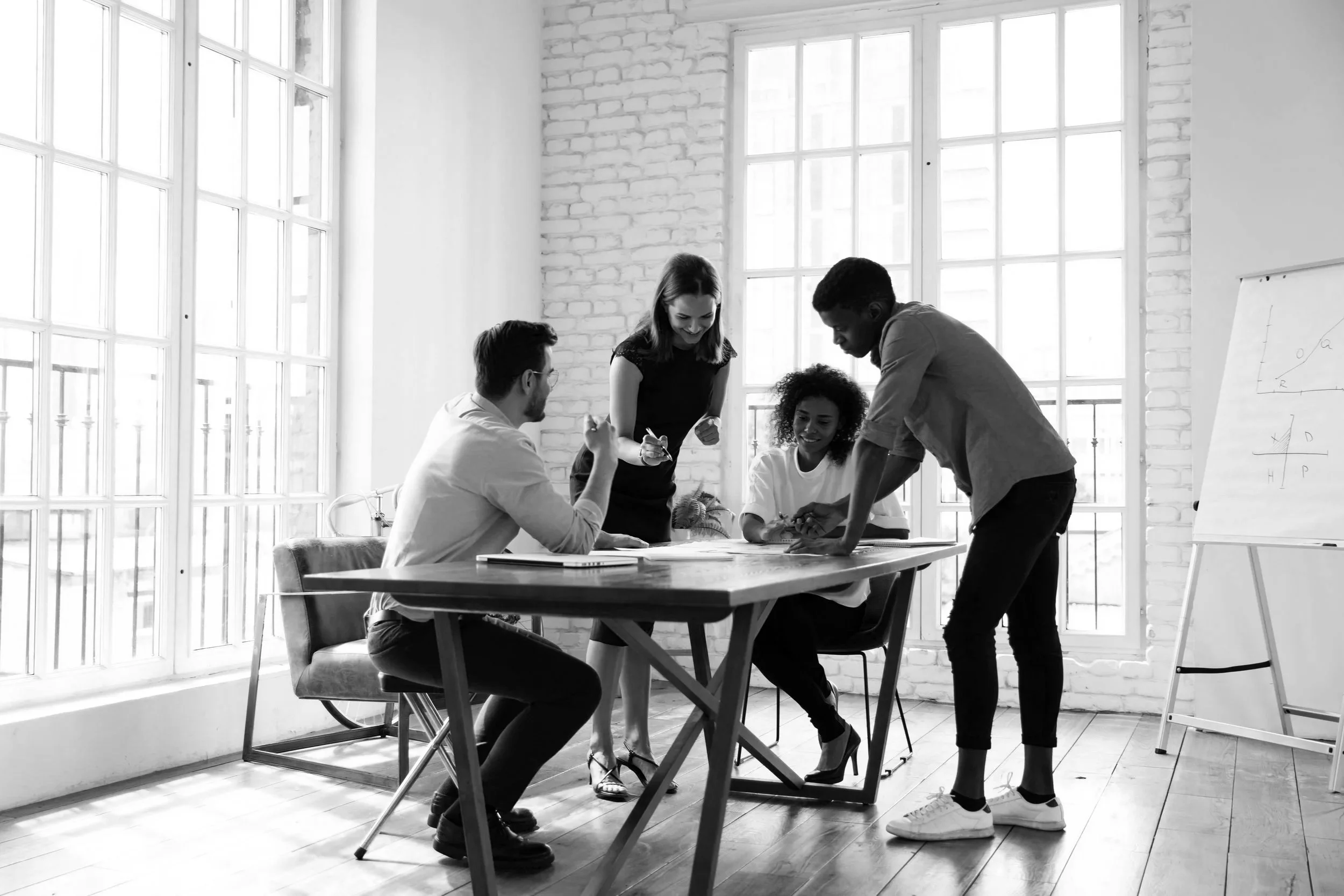 Group of five diverse people working together around a table in a modern office with large windows, white brick walls, and a whiteboard.