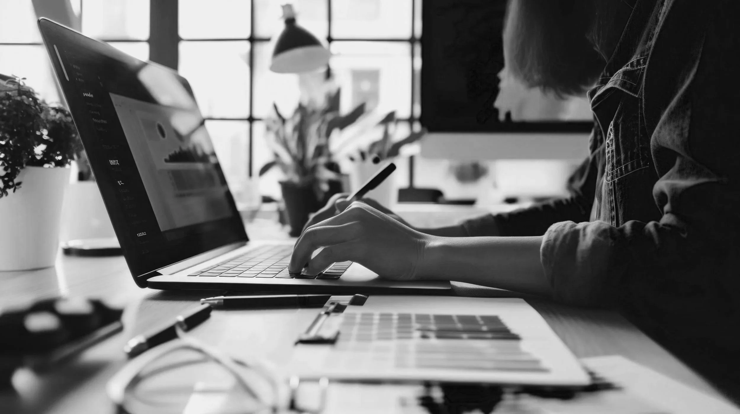 A person working on a laptop, holding a smartphone, in a well-lit room with large windows, potted plants, and several office supplies on the desk in black and white.