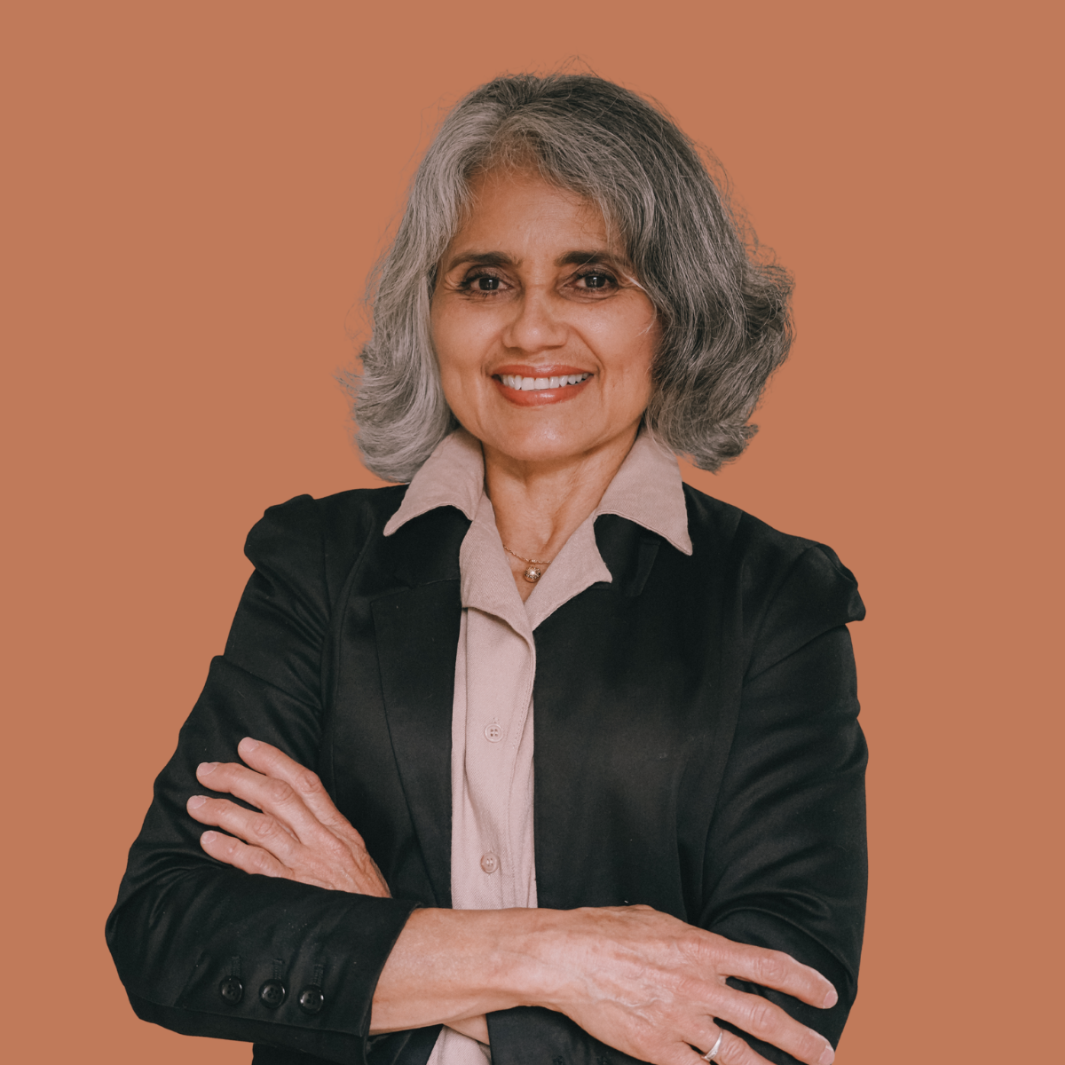 A portrait of a middle-aged woman with gray hair, smiling, wearing a black blazer over a beige collared shirt, standing against a brown background.