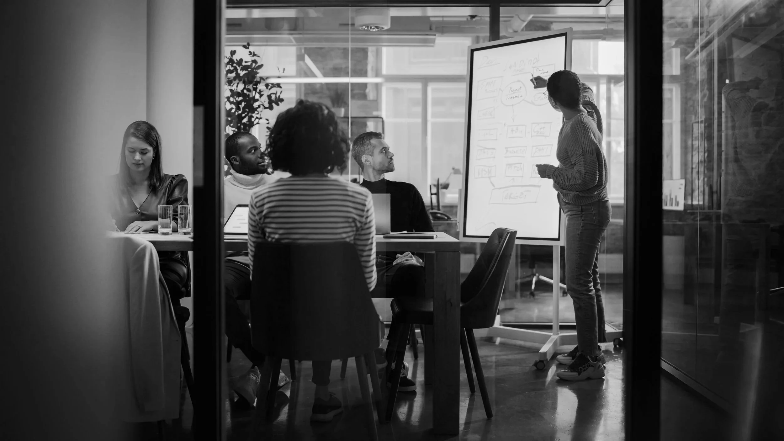 A group of five people are having a meeting or presentation in a modern office conference room. Four people are seated around a table watching a woman standing by a whiteboard, which has diagrams and notes on it.