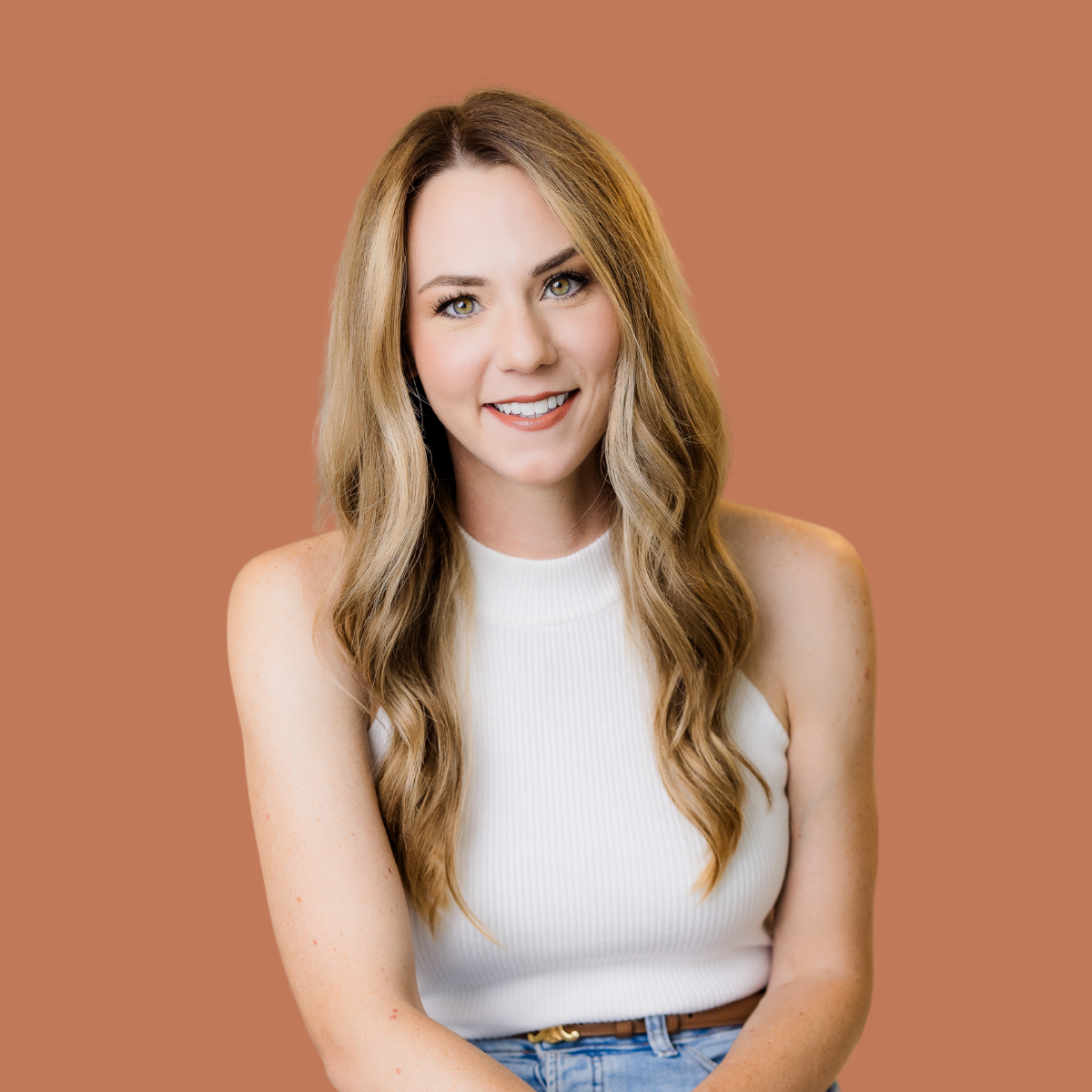 A smiling young woman with long wavy blonde hair, wearing a sleeveless white top and jeans, against a plain brown background.