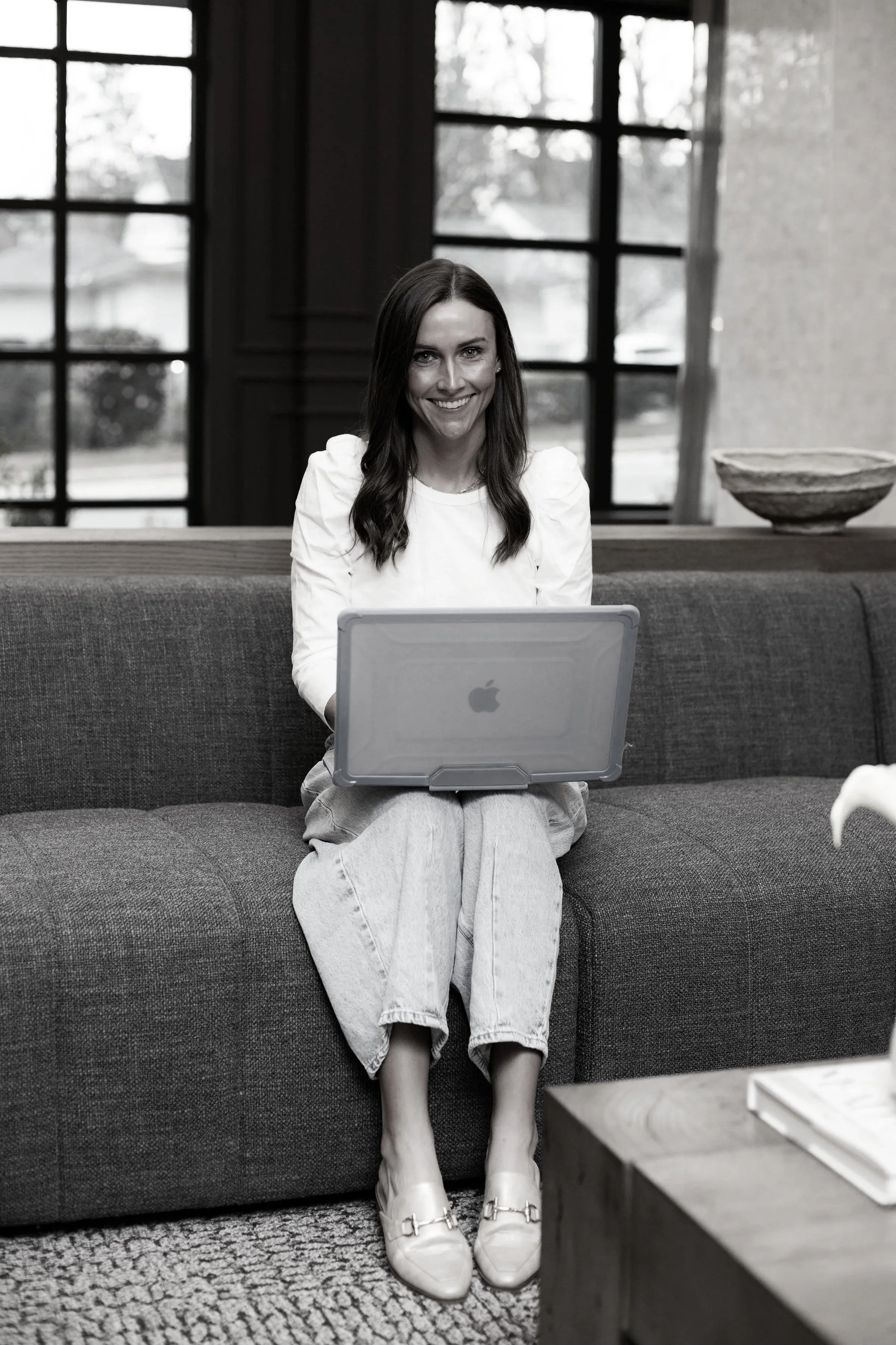 Woman sitting on a sofa with a laptop, smiling, in a room with large windows.