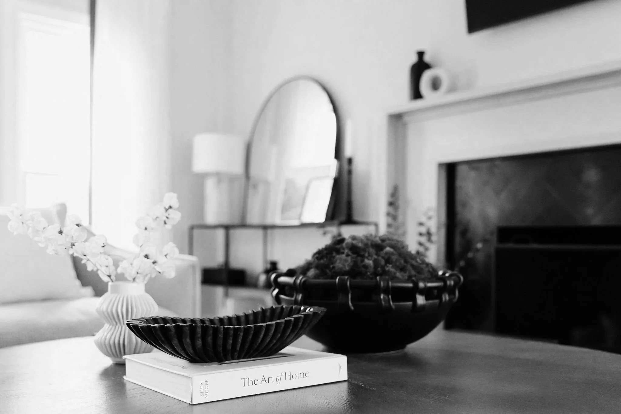 Decorative interior scene featuring a white vase with flowers, a black bowl, and a white book titled 'The Art of Home' on a wooden table, with a mirror, lamp, and fireplace in the background.