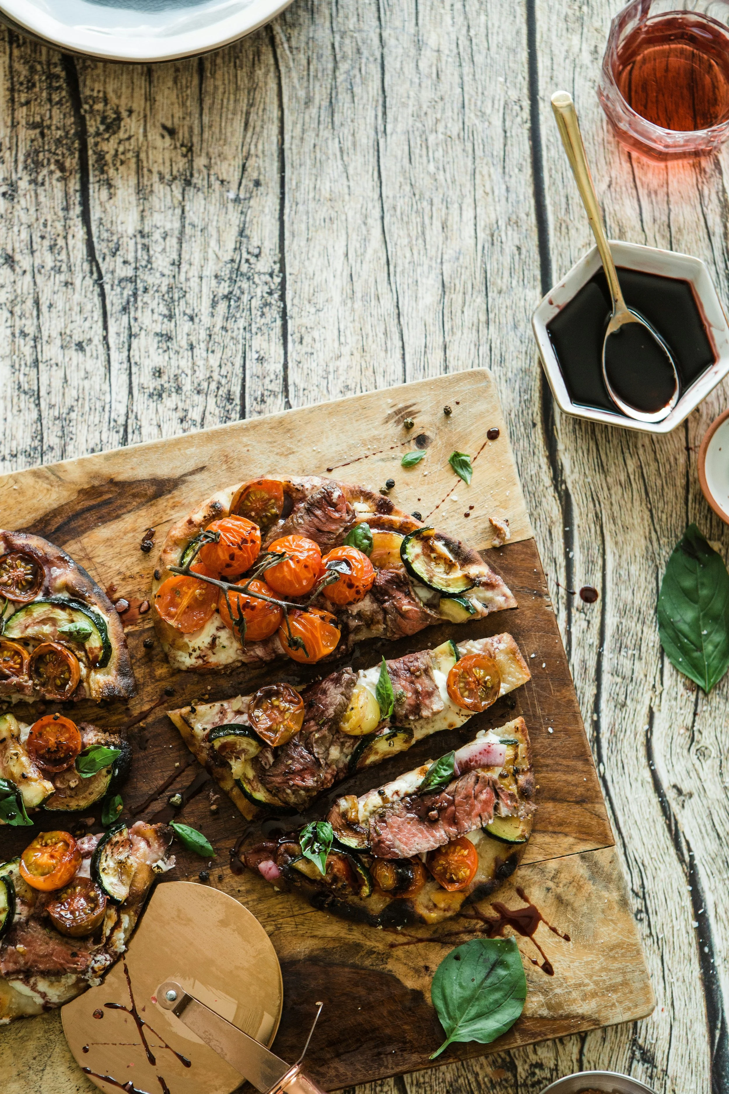 Wooden serving board with sliced gourmet pizza topped with cherry tomatoes, zucchini, and fresh basil leaves, on a rustic wooden table with a glass of rosé wine, a small bowl of balsamic vinegar, and scattered basil leaves.