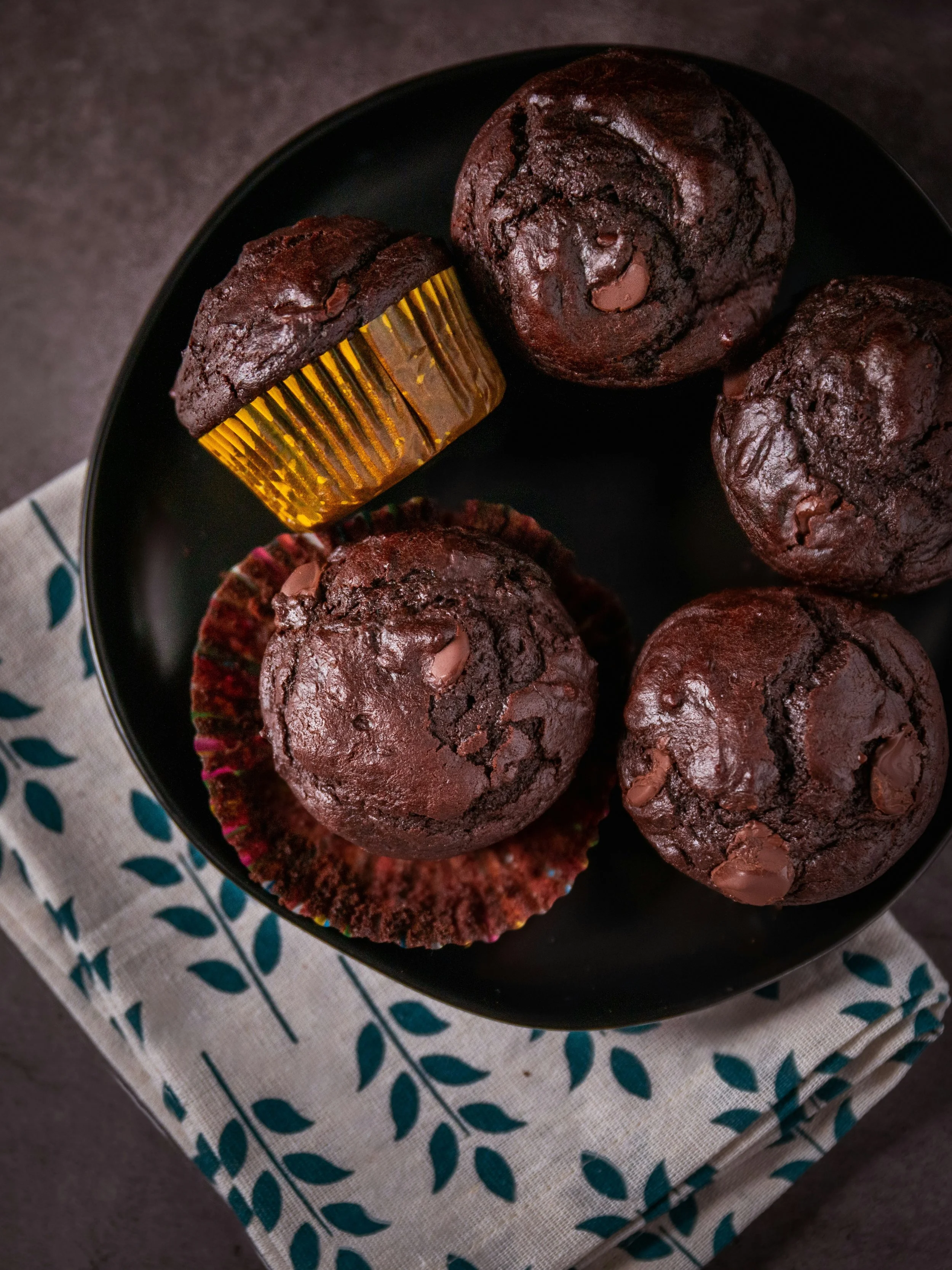 A black plate with five chocolate muffins on a white cloth with green leaf patterns, with one muffin in a colorful cupcake liner and one in a gold foil wrapper.