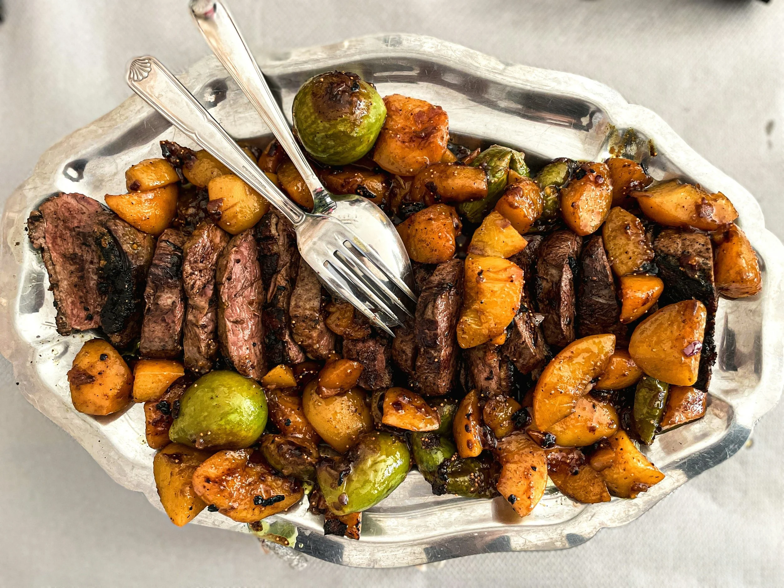 Steak slices with roasted potatoes, green olives, and cooked green peppers on a silver platter.