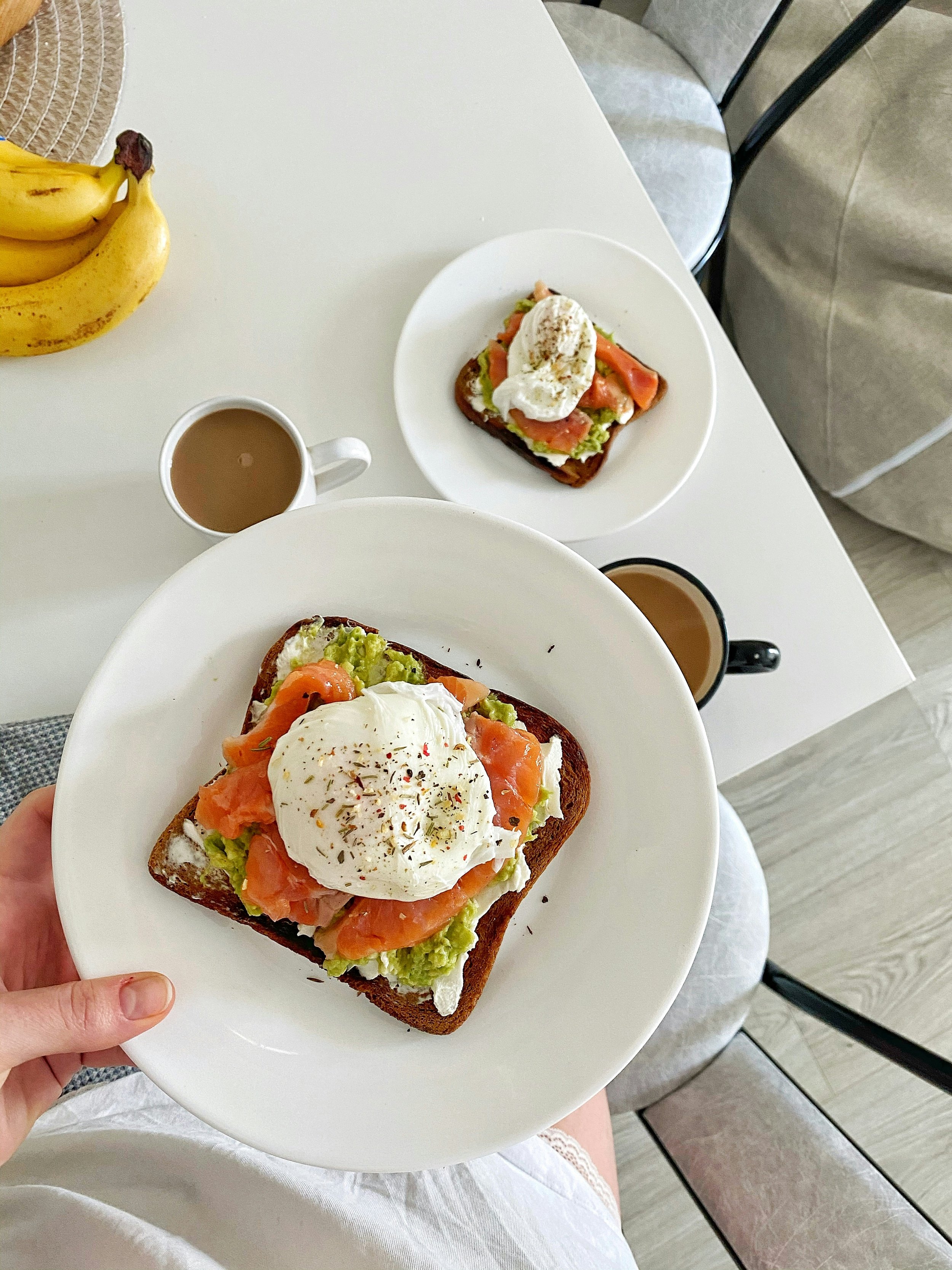 A plate of avocado toast topped with smoked salmon and a poached egg, served with a coffee. Two similar dishes are on the table, along with a bunch of bananas and a glass of coffee.