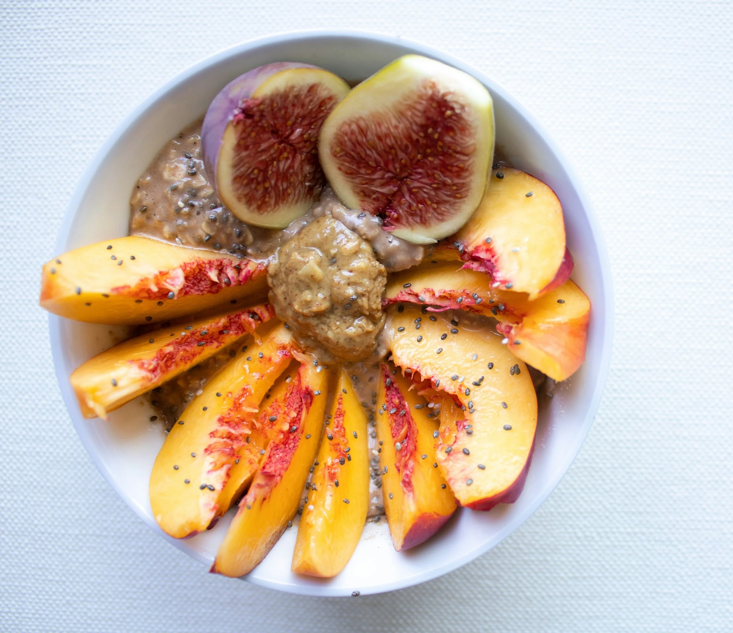A white bowl filled with sliced peaches and figs topped with chia seeds and an oat-based pudding.