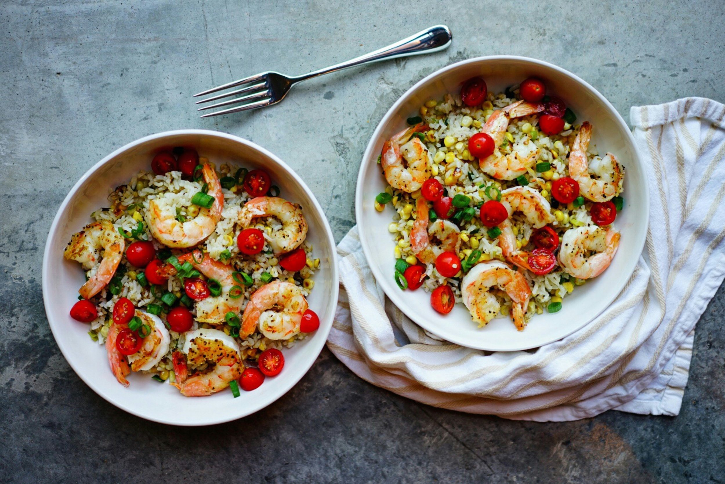 Two white plates of shrimp and rice salad with cherry tomatoes and green onions, one plate on a striped cloth, with a fork above, on a textured gray surface.