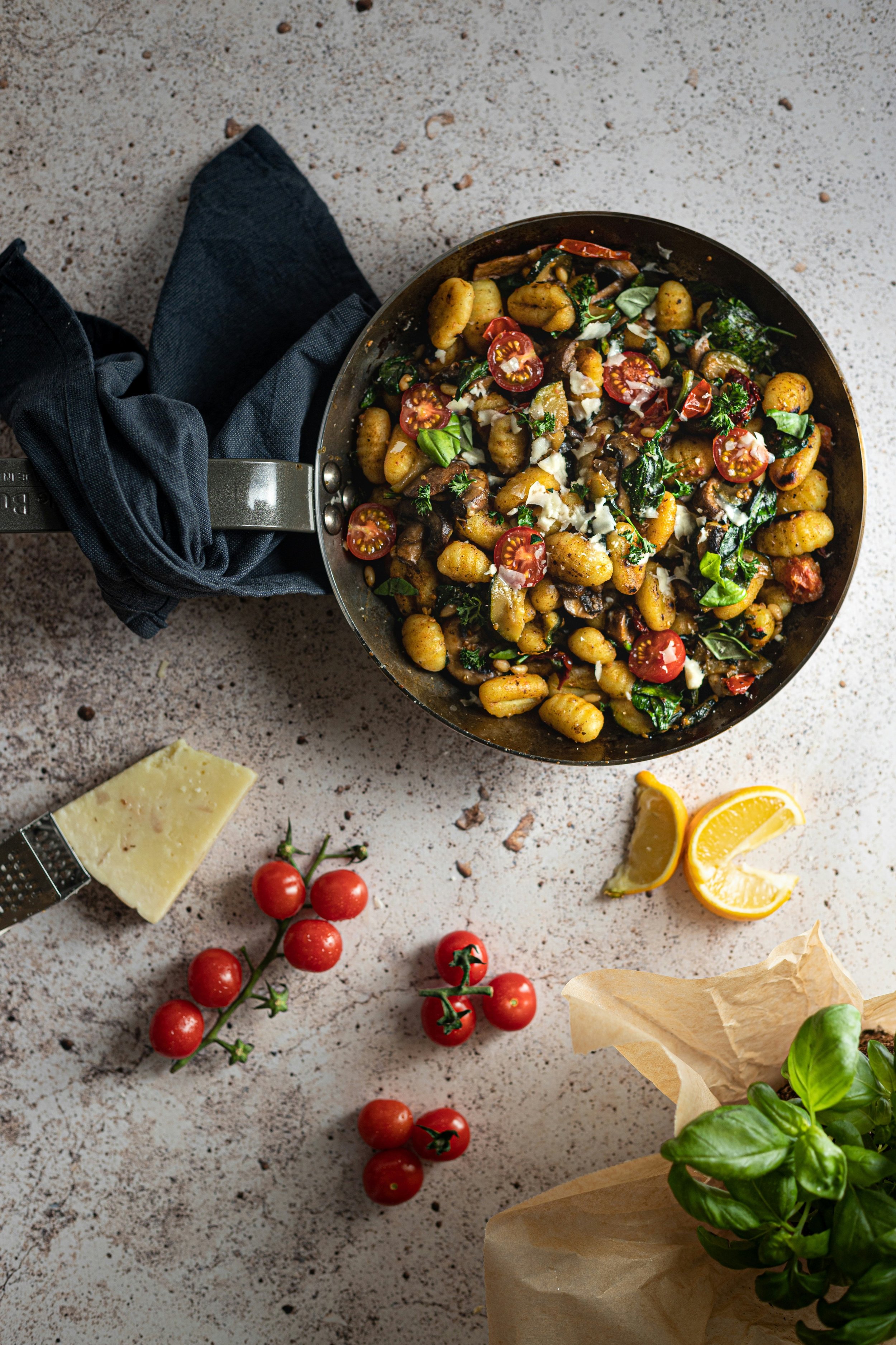 A skillet filled with gnocchi, cherry tomatoes, spinach, and cheese on a textured surface, with cherry tomatoes, cheese, lemon wedges, and basil nearby.