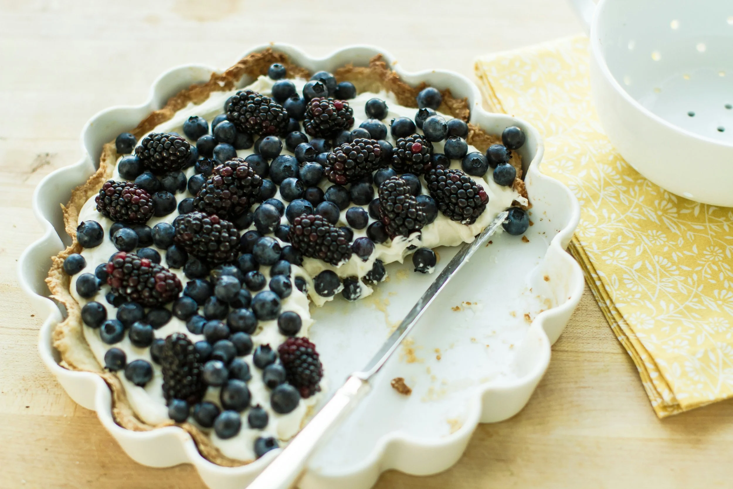 Berry tart with blueberries and blackberries in a white ceramic dish on a wooden surface, with a silver serving utensil and a yellow patterned napkin beside it.