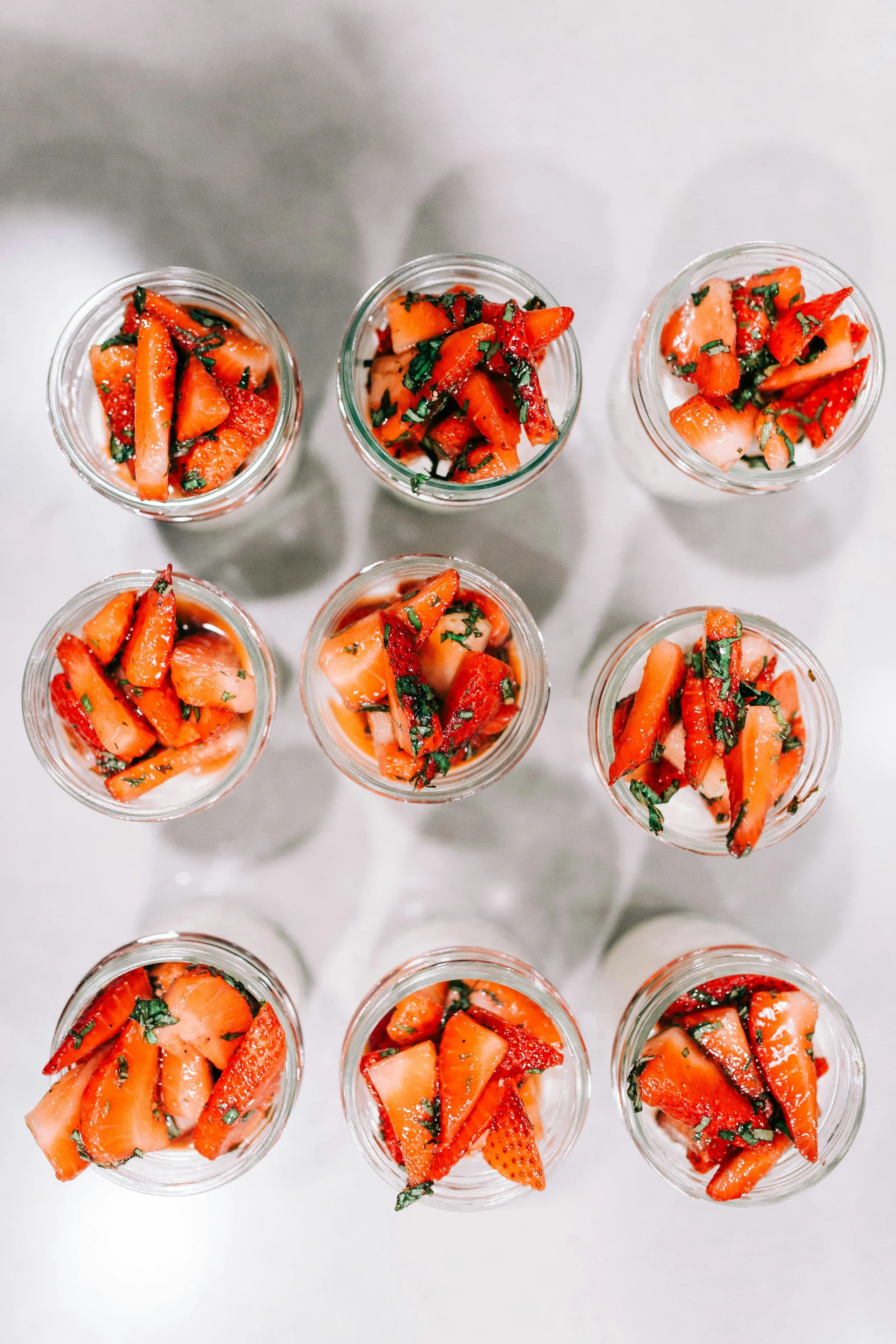Top-down view of nine small glass jars filled with chopped strawberries and fresh herbs, arranged on a white surface.