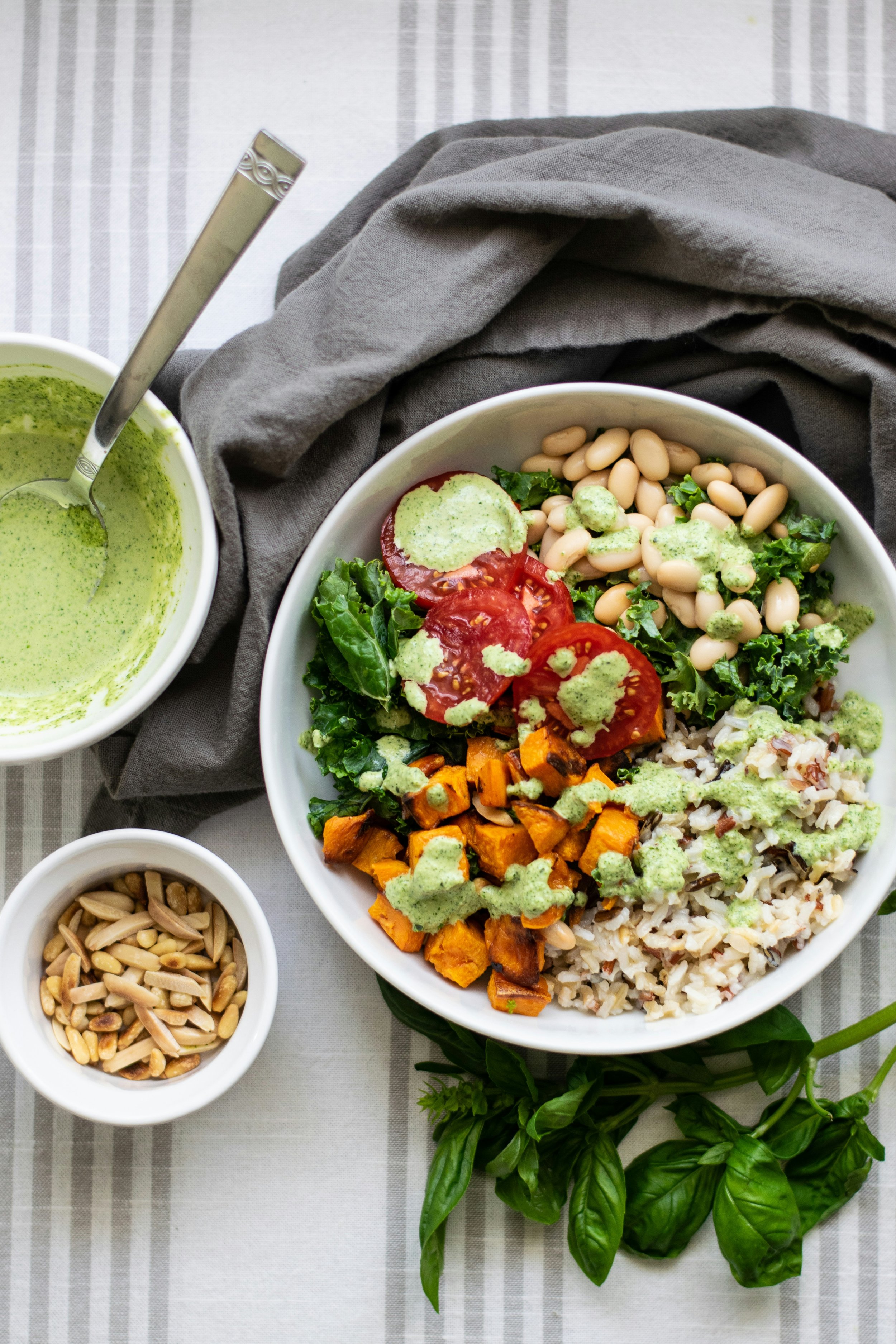 A bowl of salad with tomatoes, kale, roasted sweet potatoes, rice, and dressing, with a side of white beans, a small bowl of sunflower seeds, and a cup with green dressing, placed on a table with a gray cloth and a bunch of fresh basil.