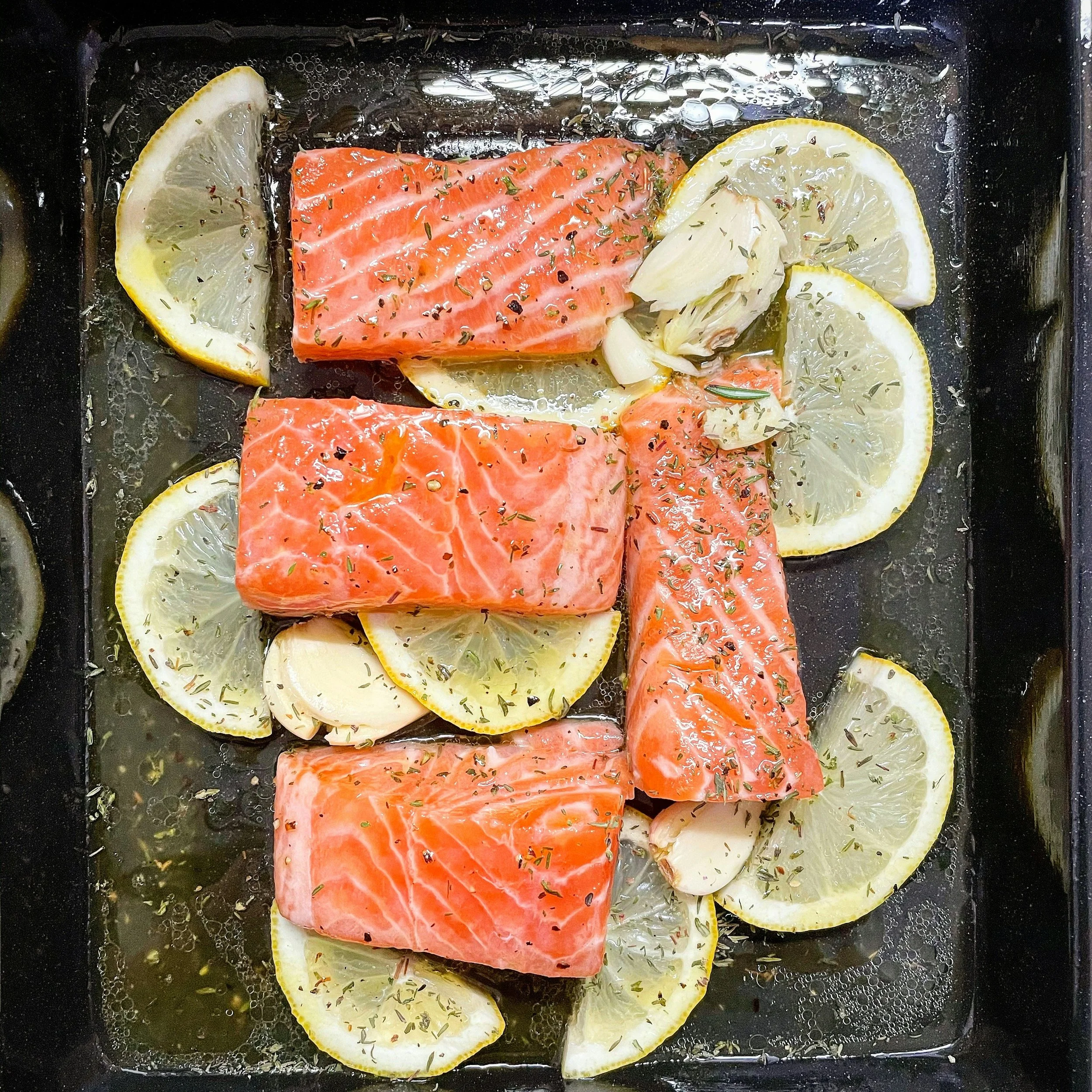 Raw salmon fillets seasoned with herbs on a baking tray surrounded by lemon slices and garlic cloves.