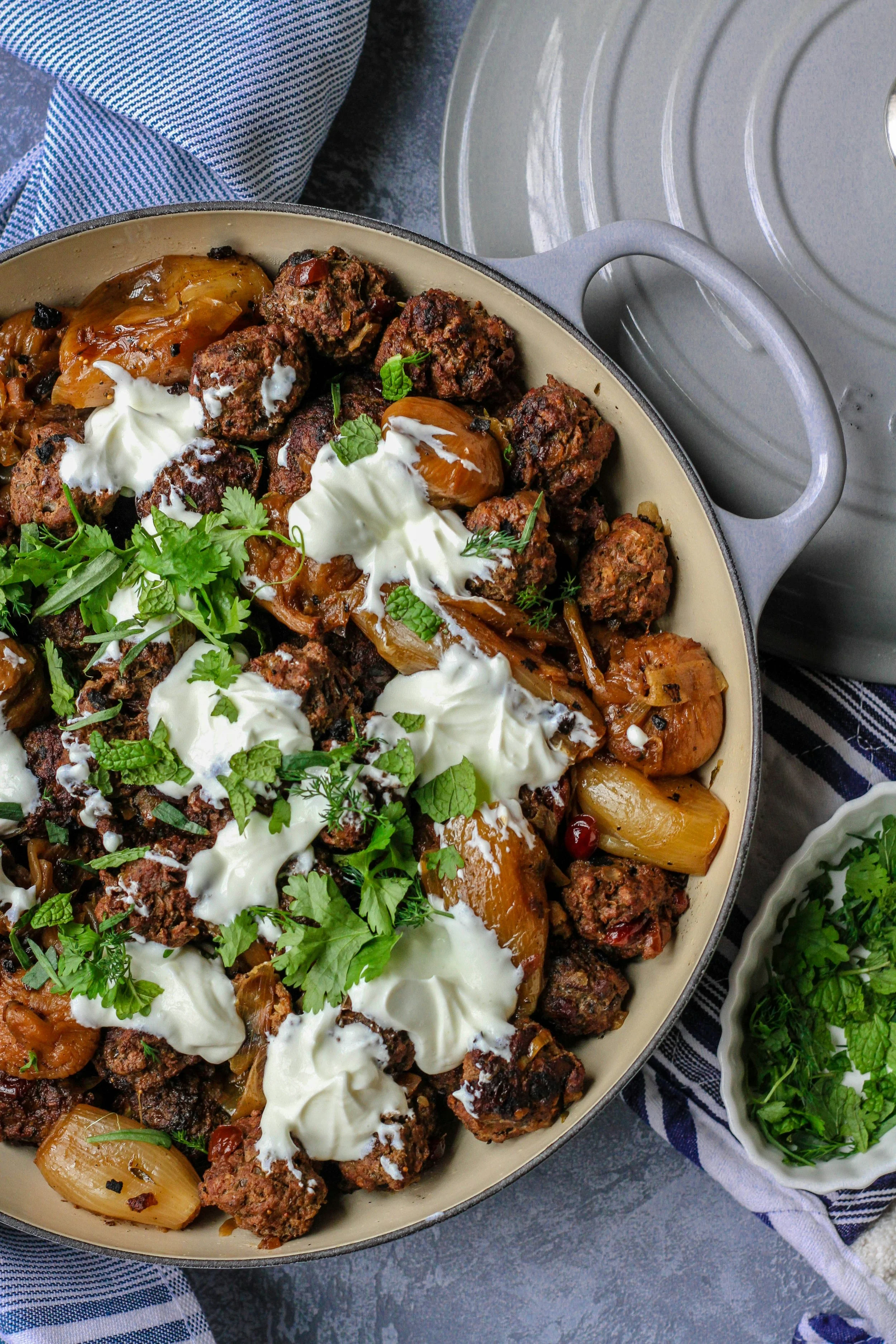 A skillet filled with cooked meatballs, caramelized onions, and sauce, garnished with fresh herbs and a dollop of sour cream. A small bowl of chopped herbs is beside it.