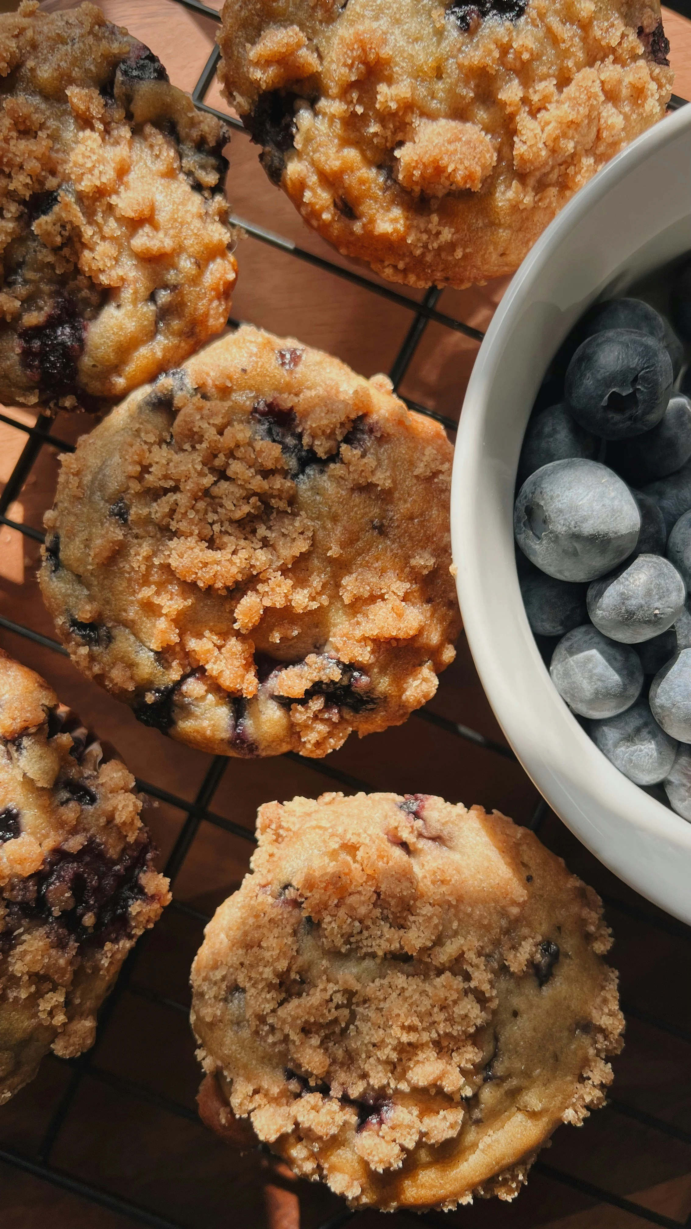 Close-up of blueberry muffins with a crumbly topping, set on a cooling rack, with a bowl of fresh blueberries nearby.