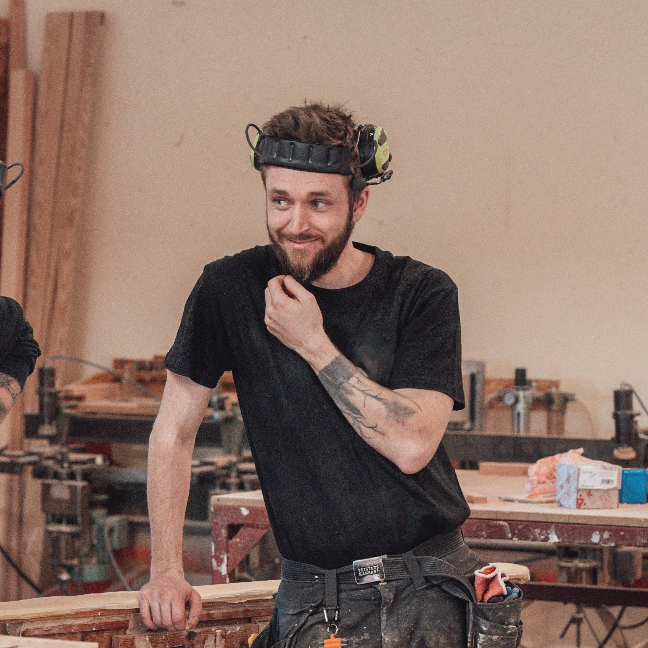 Man i svart T-shirt, med verktyg i bältesfästet, ler i ett snickeri, med hörselskydd på huvudet, omgiven av trä och verktyg.