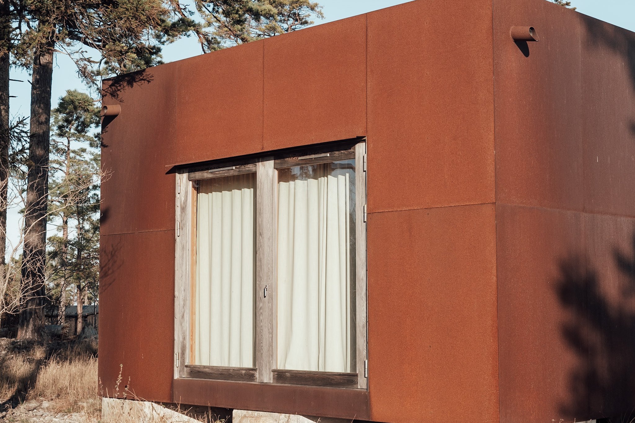 Ett modernt, rustfärgat hus med stora fönster och trädetallar i ett skogig landskap