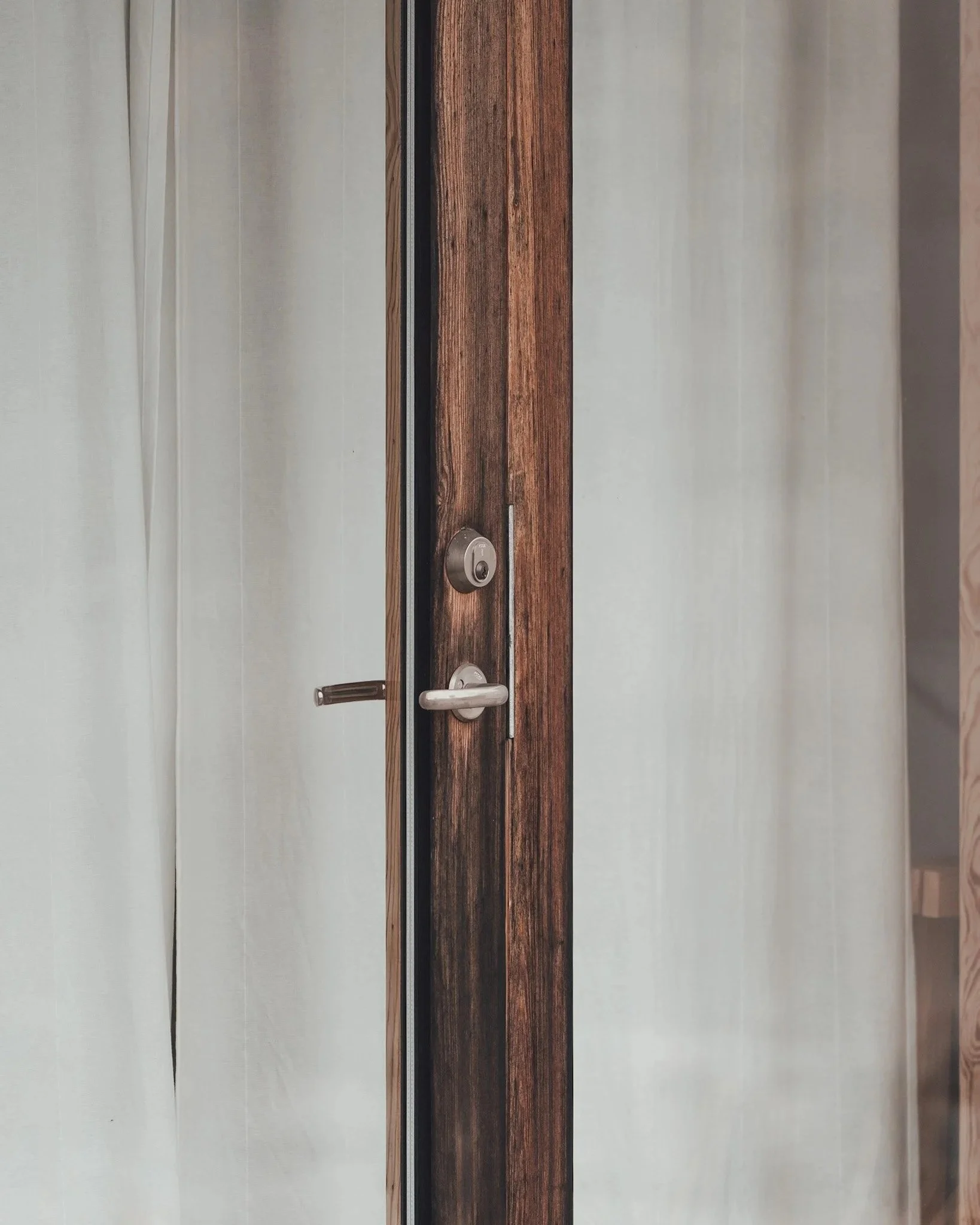 Dörr med träkarm, lås och handtag, invändigt vita gardiner som hänger på sidan.