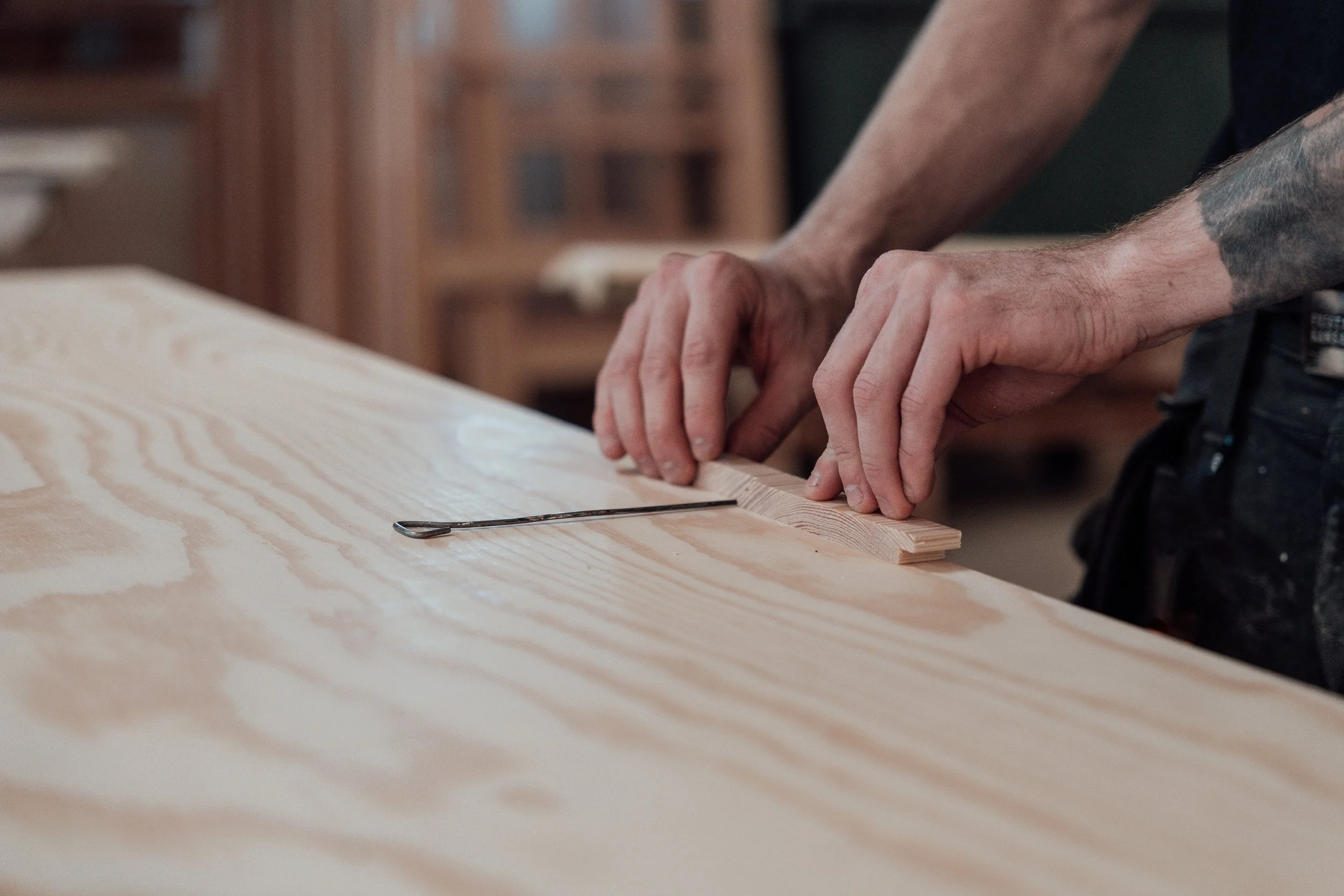 Två personer arbetar tillsammans i ett träslöjdsverkstad, där de slipar och formar träplankor. Fokus på deras händer och träskivorna.