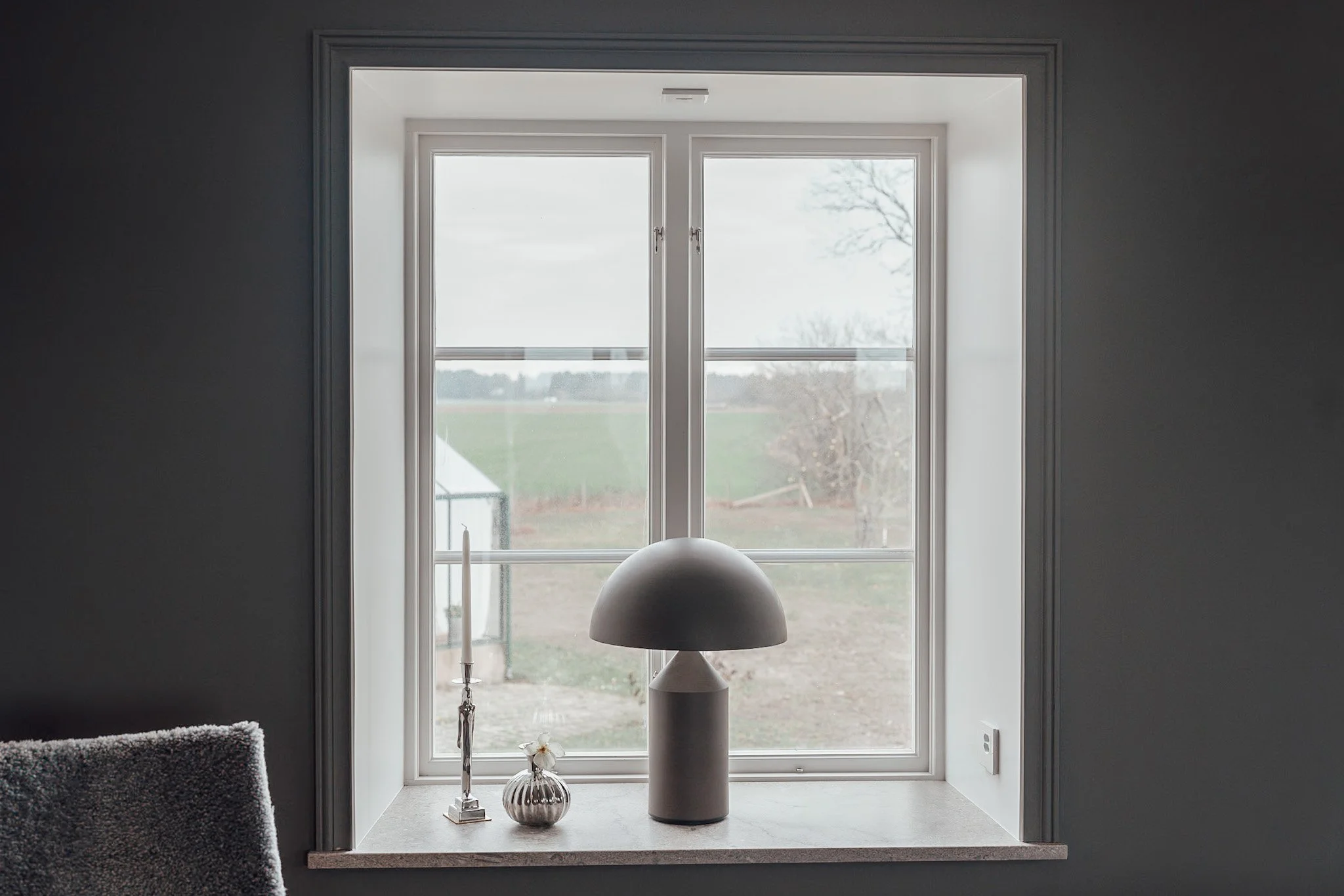 En fönsterkarm med en bordslampa, ett ljusstake och en liten blomkruka, utanför syns ett öppet fält och träd.