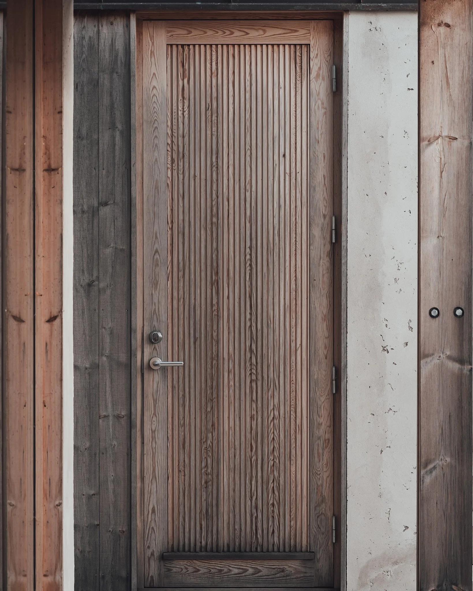 En trädörr med vertikala ribbor, omgiven av träväggar och en betongvägg på höger sida.