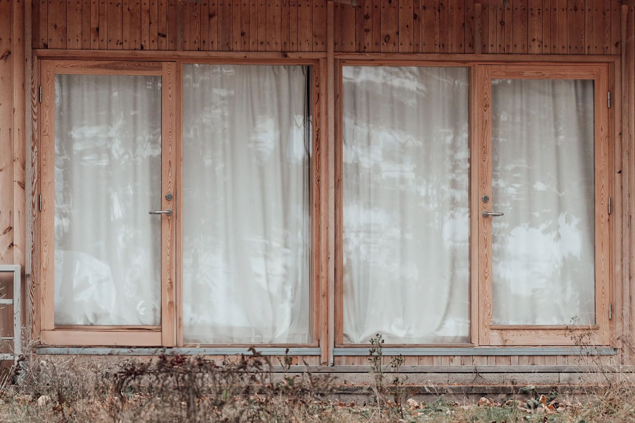Tomma glasdörrar med vita gardiner på en träfasad, med viss växtlighet och höstlöv framför.