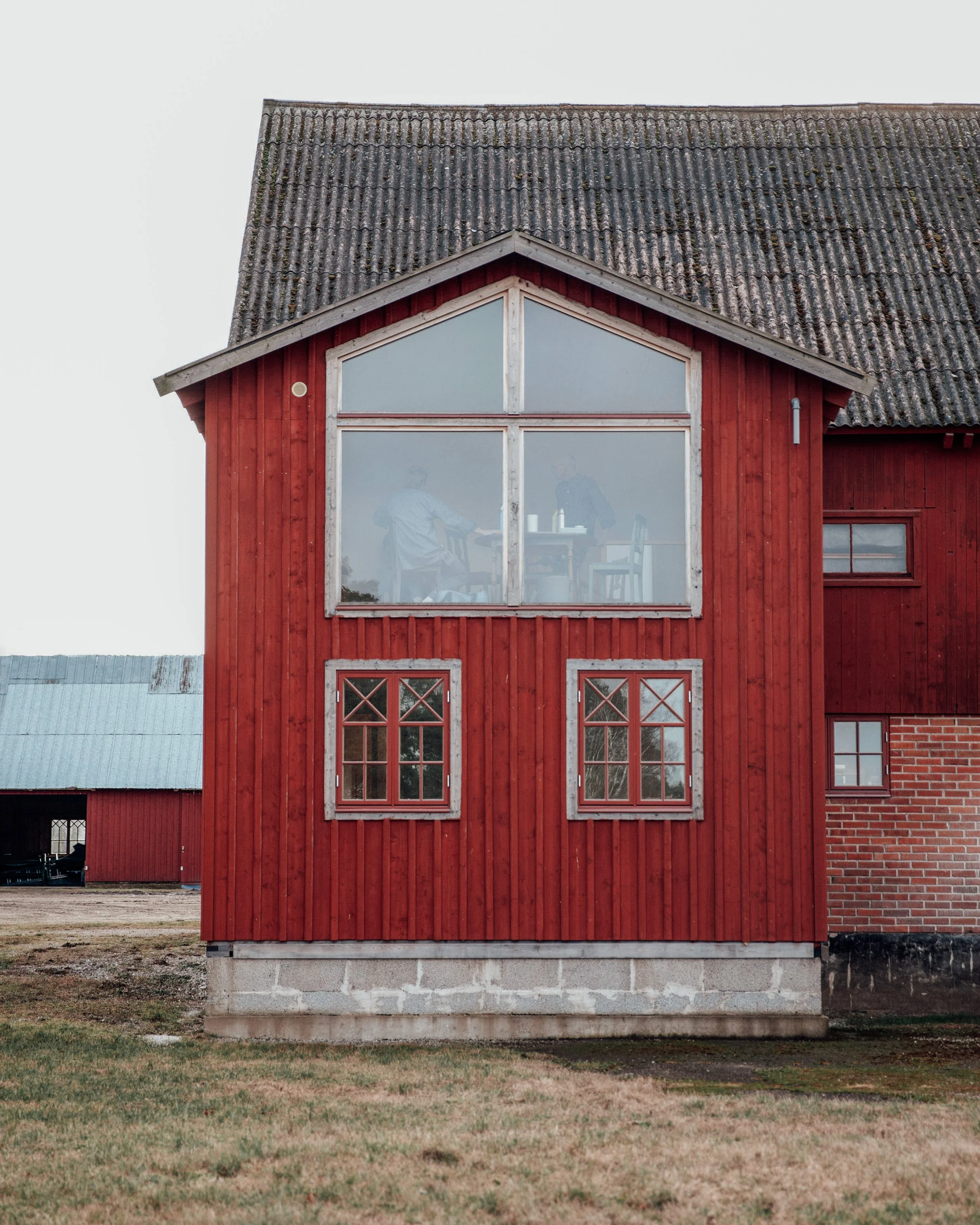 En röd trävilla med stora fönster och ett litet fönster på sidan. Det finns två personer synliga genom det övre fönstret, som sitter vid ett bord.
