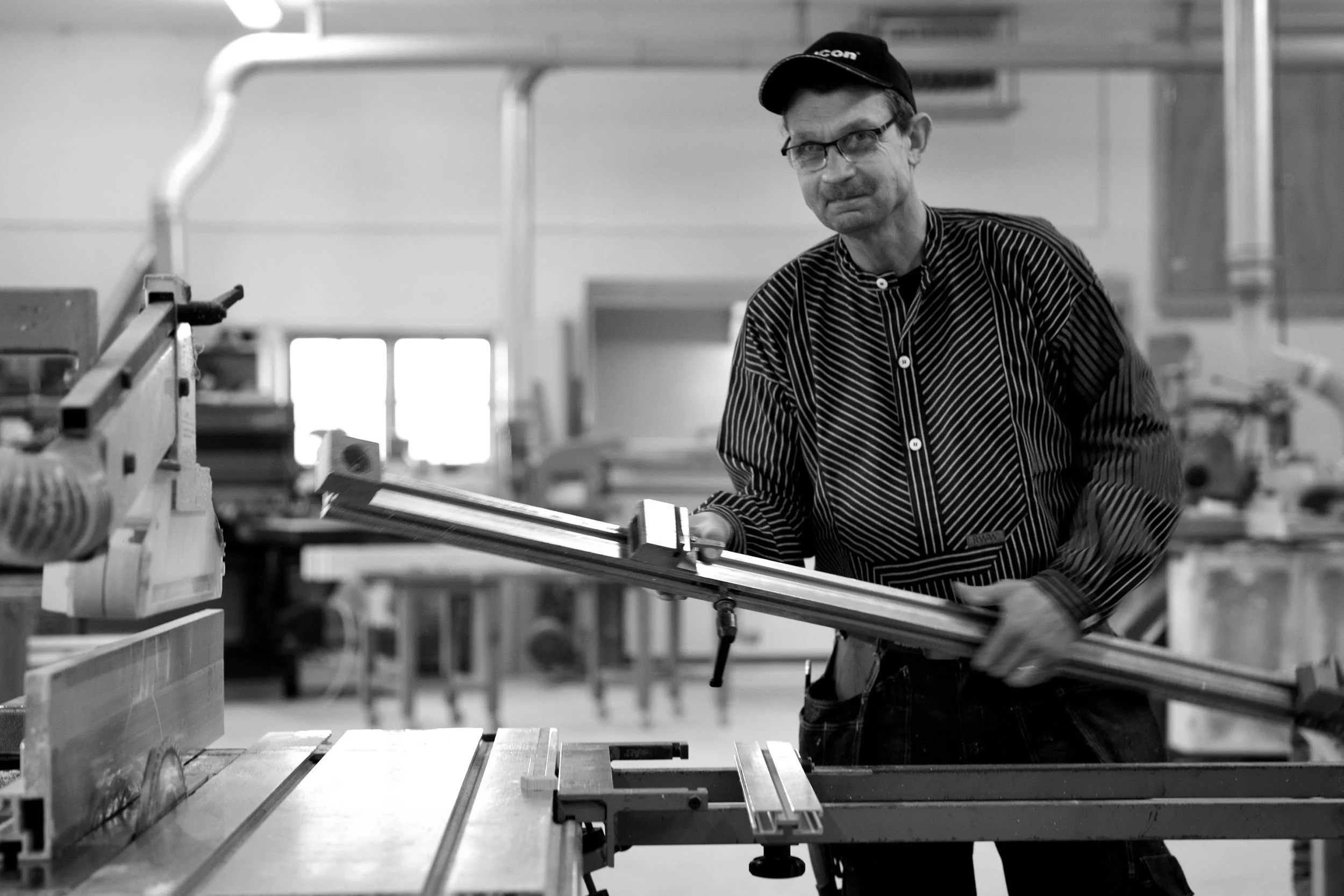En man i glasögon och keps arbetar i ett verkstadsmiljö och håller en lång metallstång.