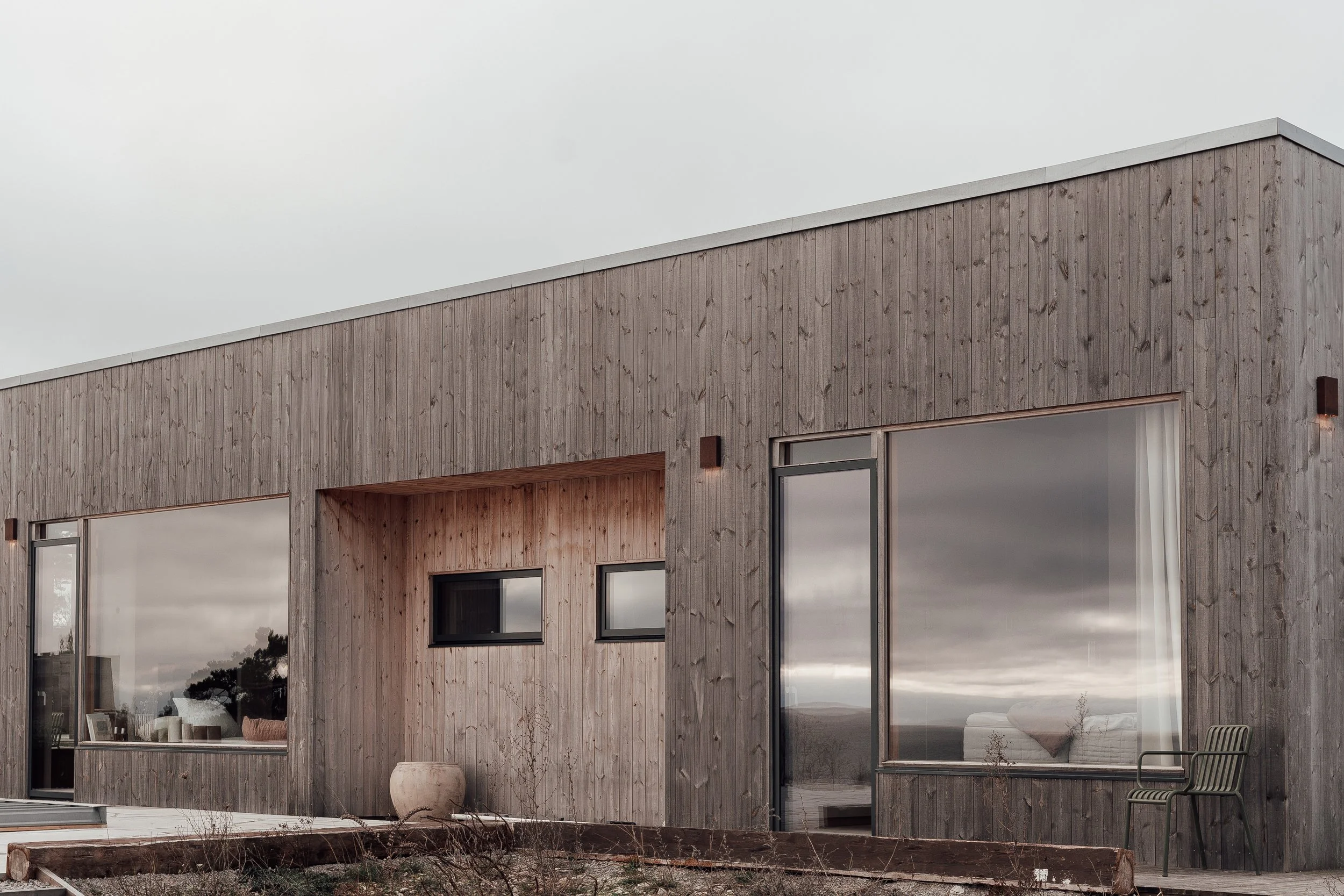 Modern trähus med stora fönster och sittplats på trädäck.