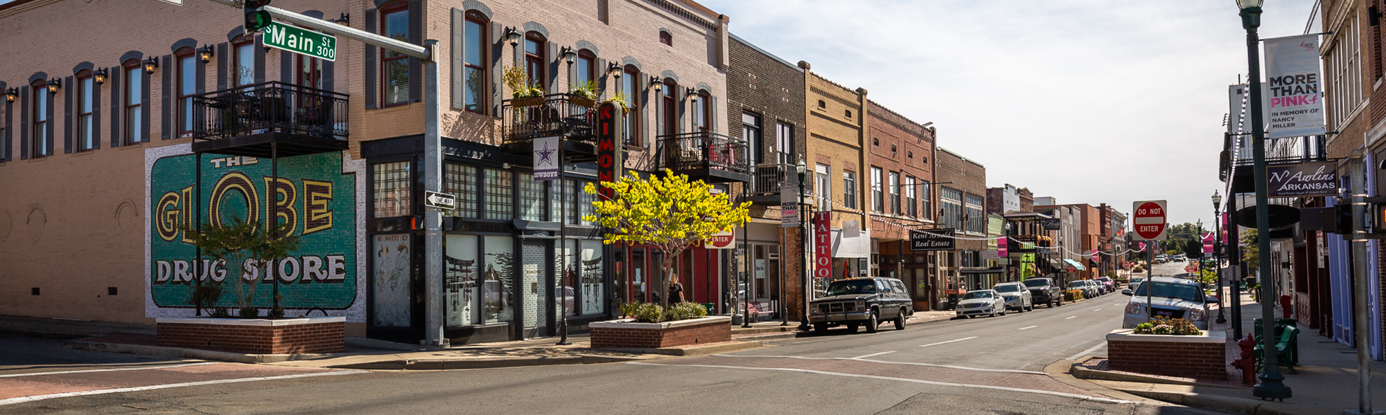 A small town main street with brick buildings, parked cars, and signs for a tattoo shop, a movie theater, and a drug store. There are lampposts, a few trees, and pink banners hanging across the street. The street signs read Main Street and one-way.