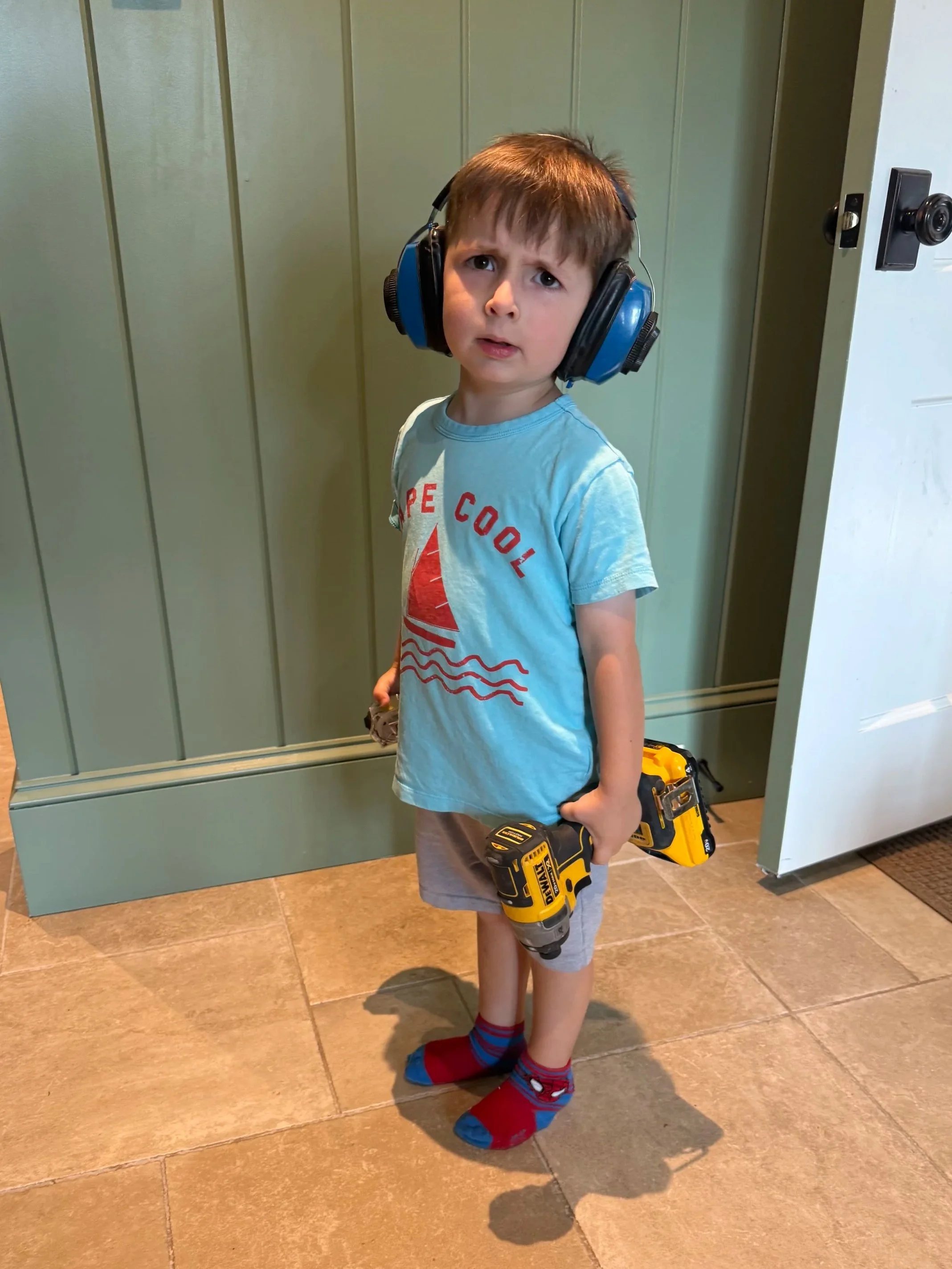 A young boy with brown hair wearing large blue safety headphones, a light blue T-shirt with a red sailboat and the words "Hope Cool," gray shorts, colorful socks, and holding two yellow and black cordless drills, standing on a brown tiled floor inside a house.