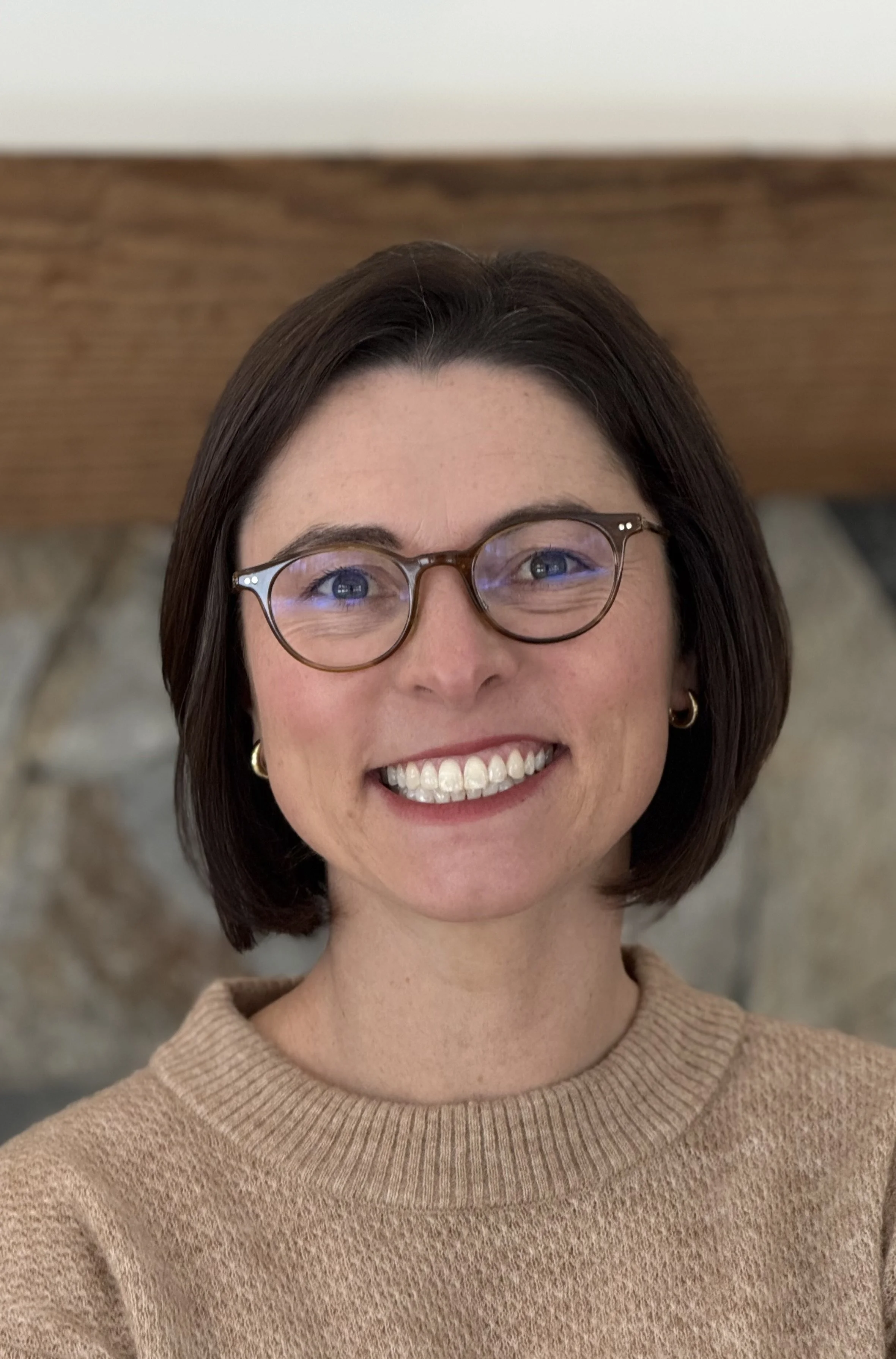 Close-up of a smiling woman with short brown hair, glasses, earrings, and a beige sweater, indoors with a stone fireplace and wooden mantle in the background.