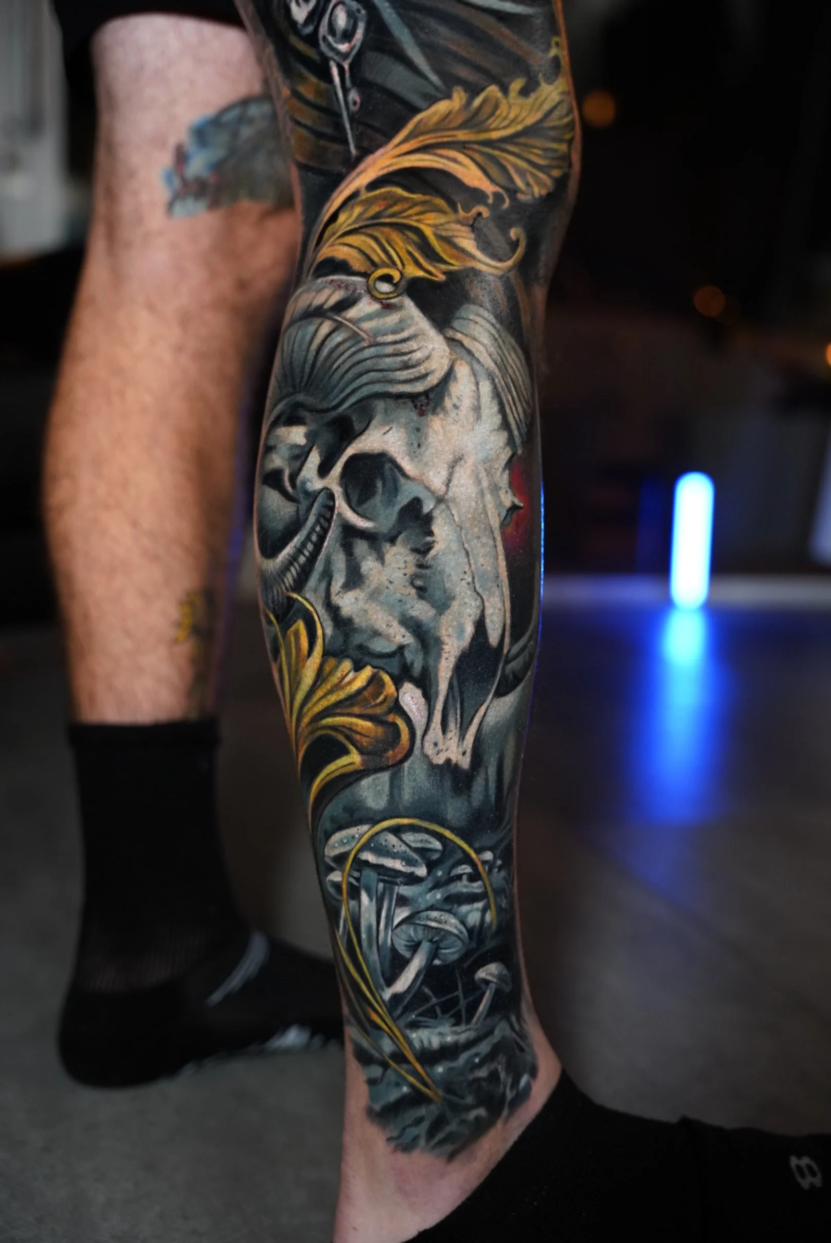 Black and grey realism calf tattoo featuring skull, mushrooms, and ornamental gold accents