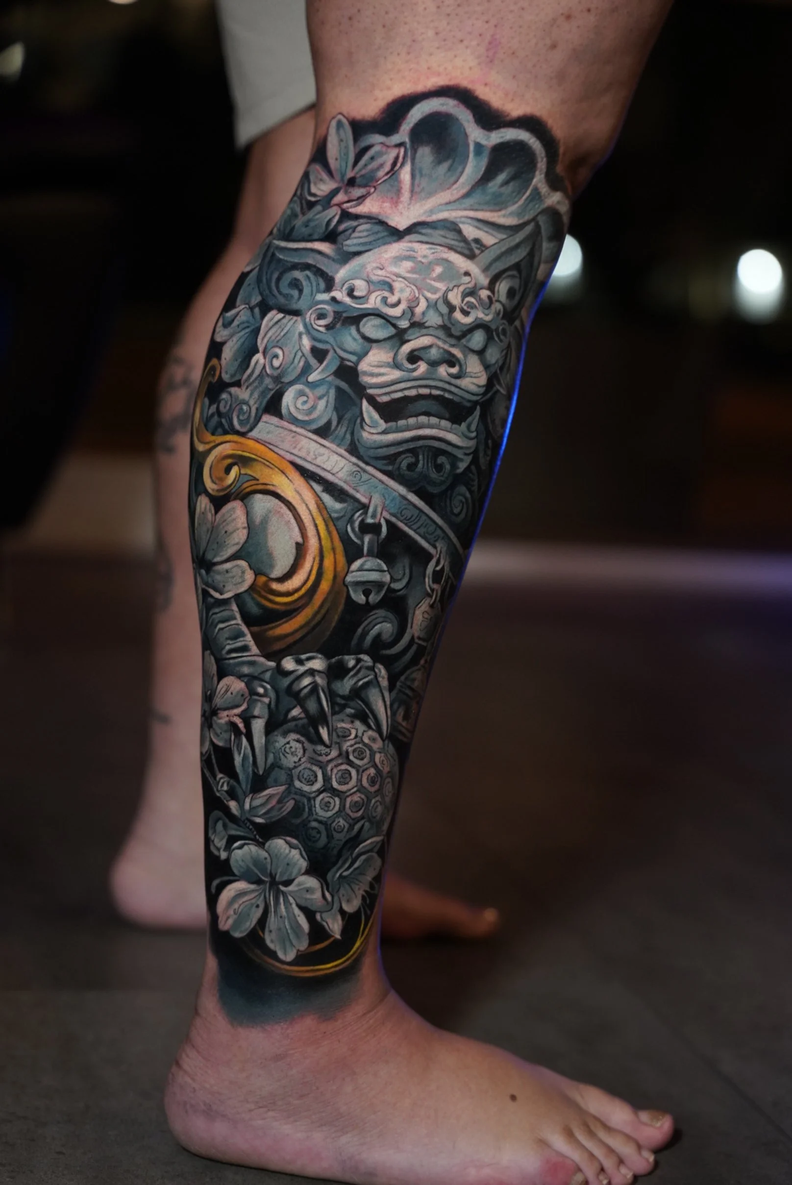 Japanese inspired black and grey realism leg tattoo featuring guardian lion mask and floral elements