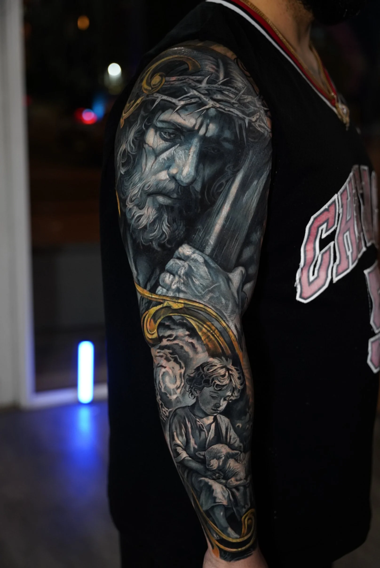 Large-scale black and grey realism tattoo featuring a symbolic composition of Christ and a child, connected through flowing ornamental elements. Part of a custom sleeve project focused on storytelling, depth, and emotional contrast.