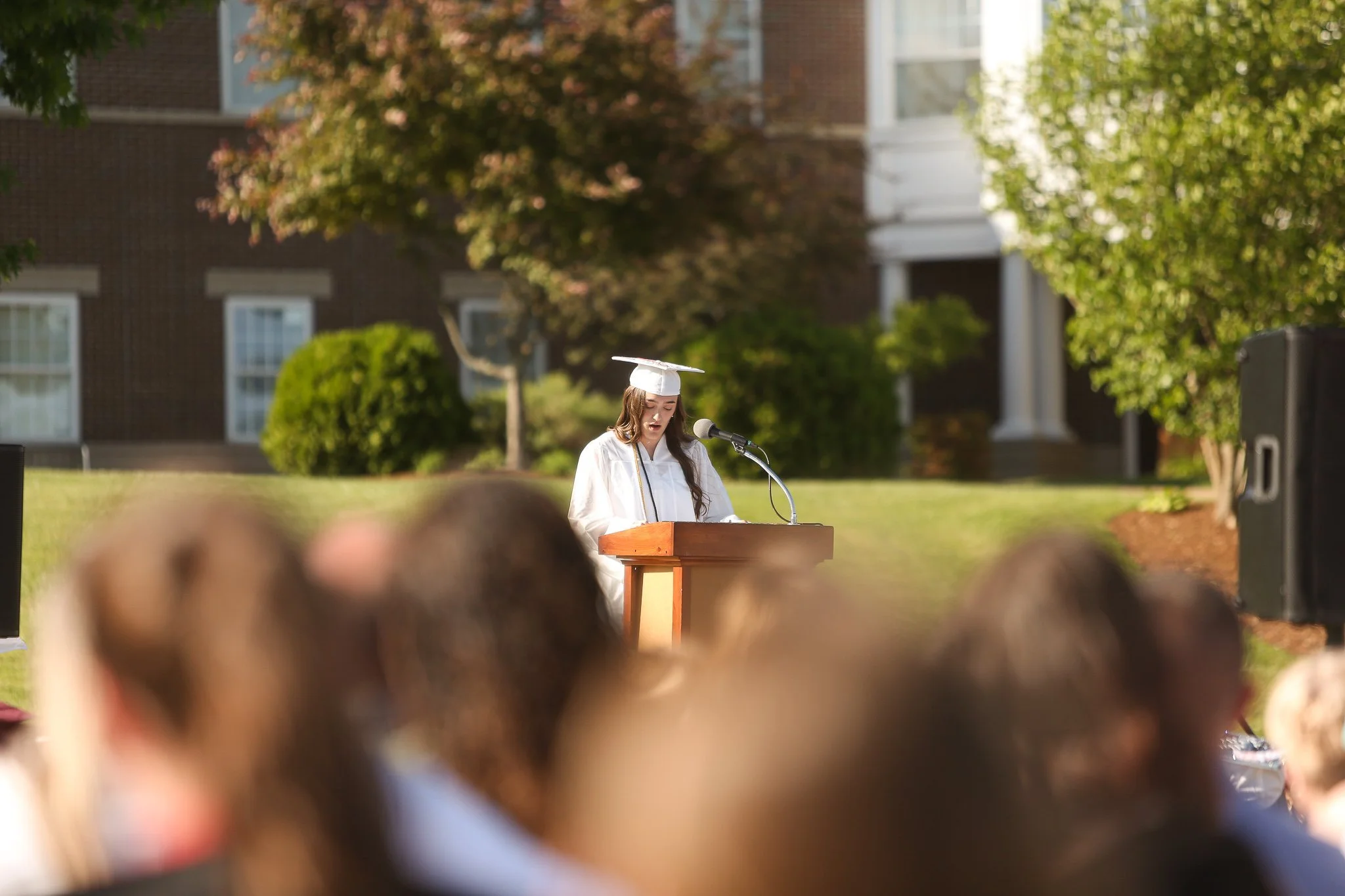 A young woman in graduation cap and gown speaking at a podium outdoors during a graduation ceremony, with a blurred audience in the foreground and a school building and trees in the background.