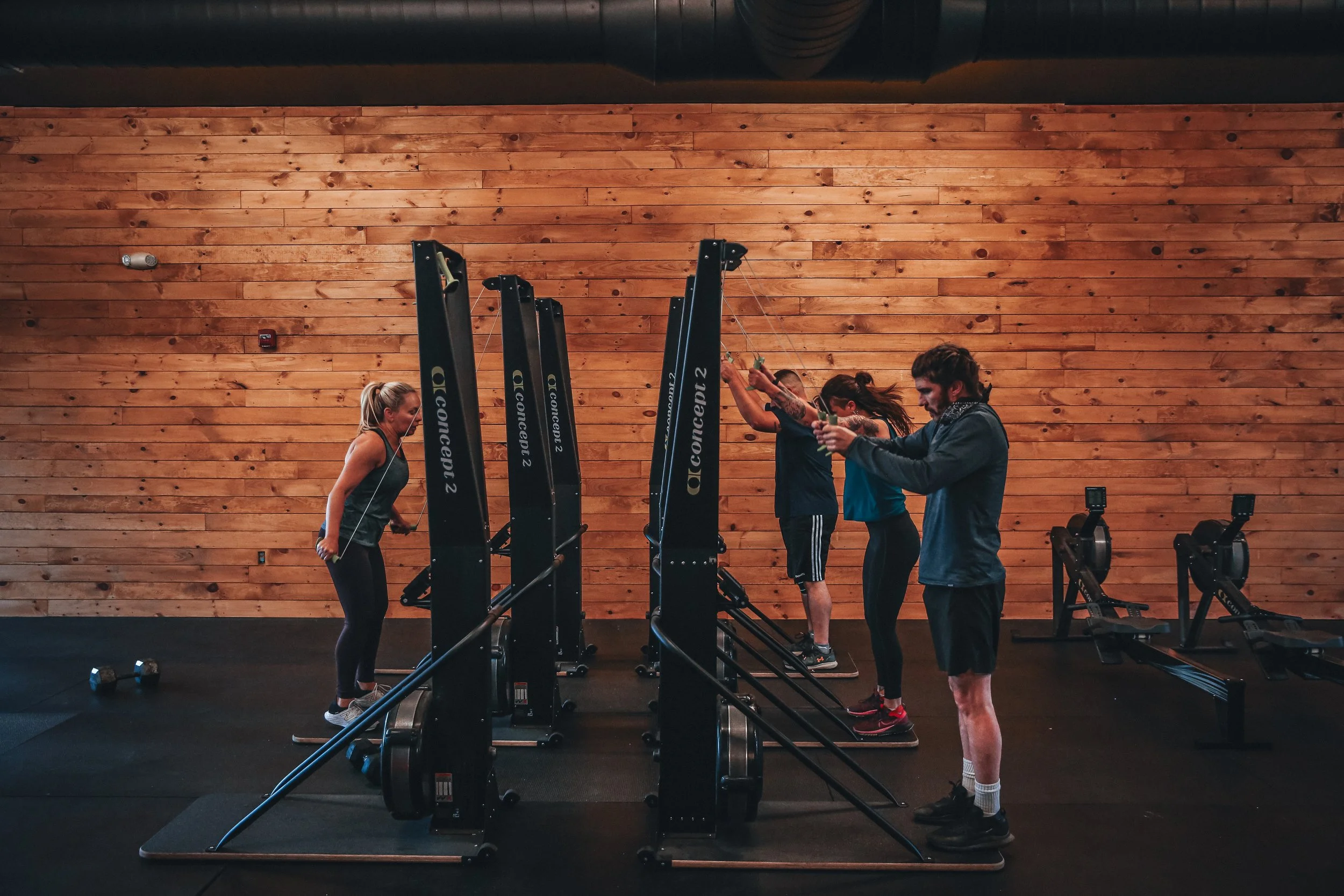 Five people using rowing machines in a gym with wooden walls.