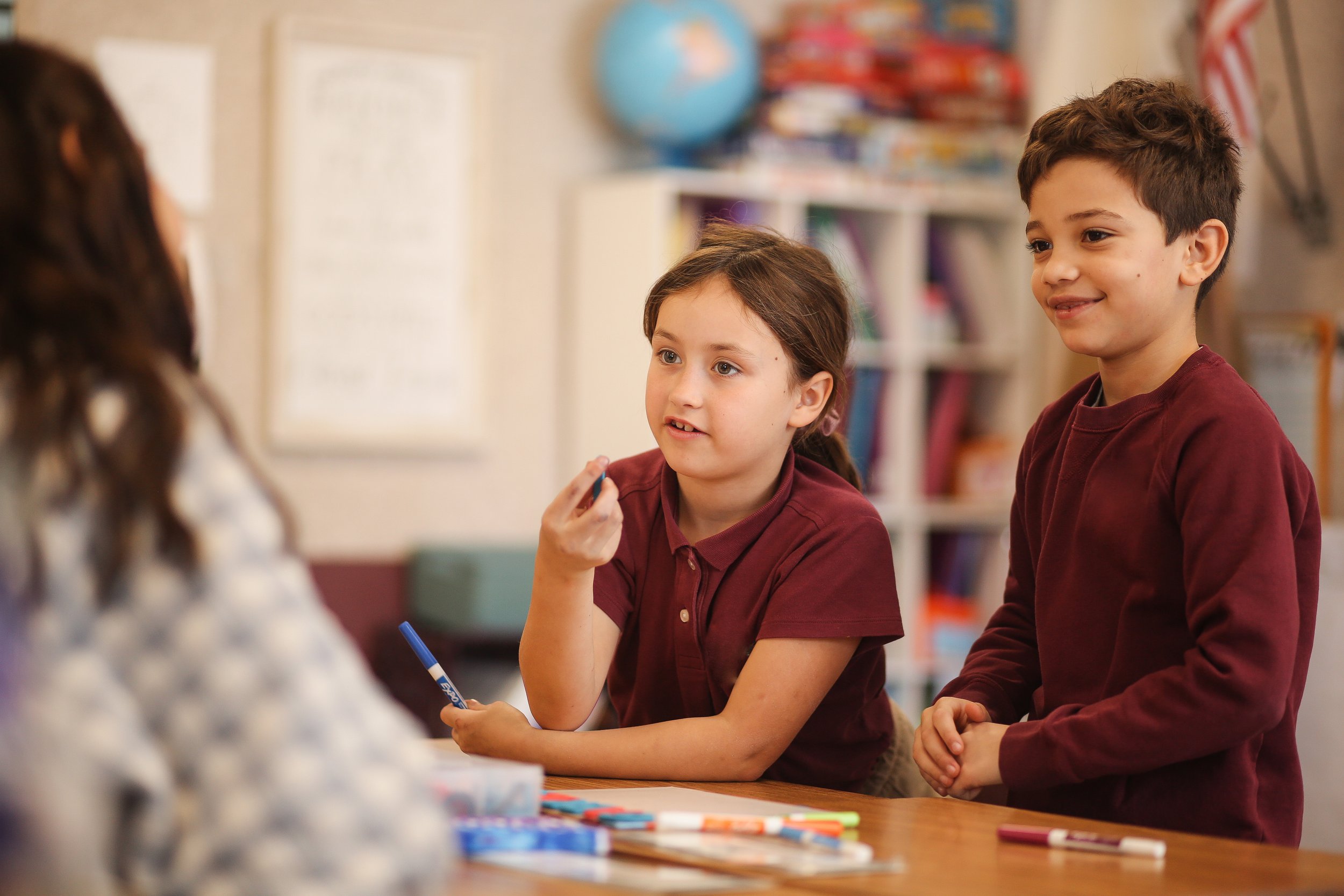 Two children, a girl and a boy, talking to a girl sitting at a table in a classroom.