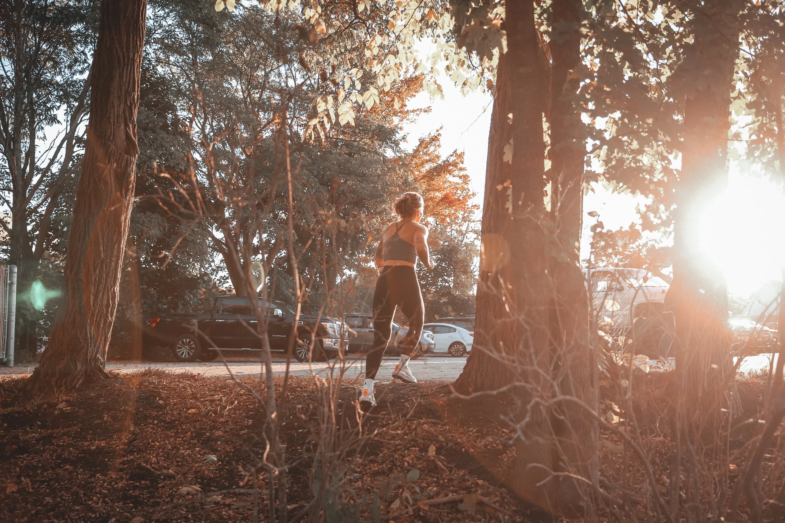 A woman running in a wooded outdoor area during sunset with parked cars in the background.