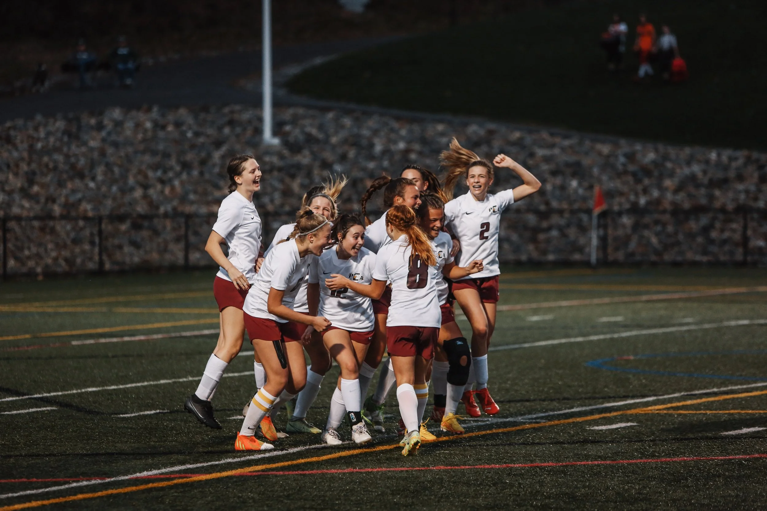A group of female soccer players in white jerseys and maroon shorts celebrating on the field during a game.