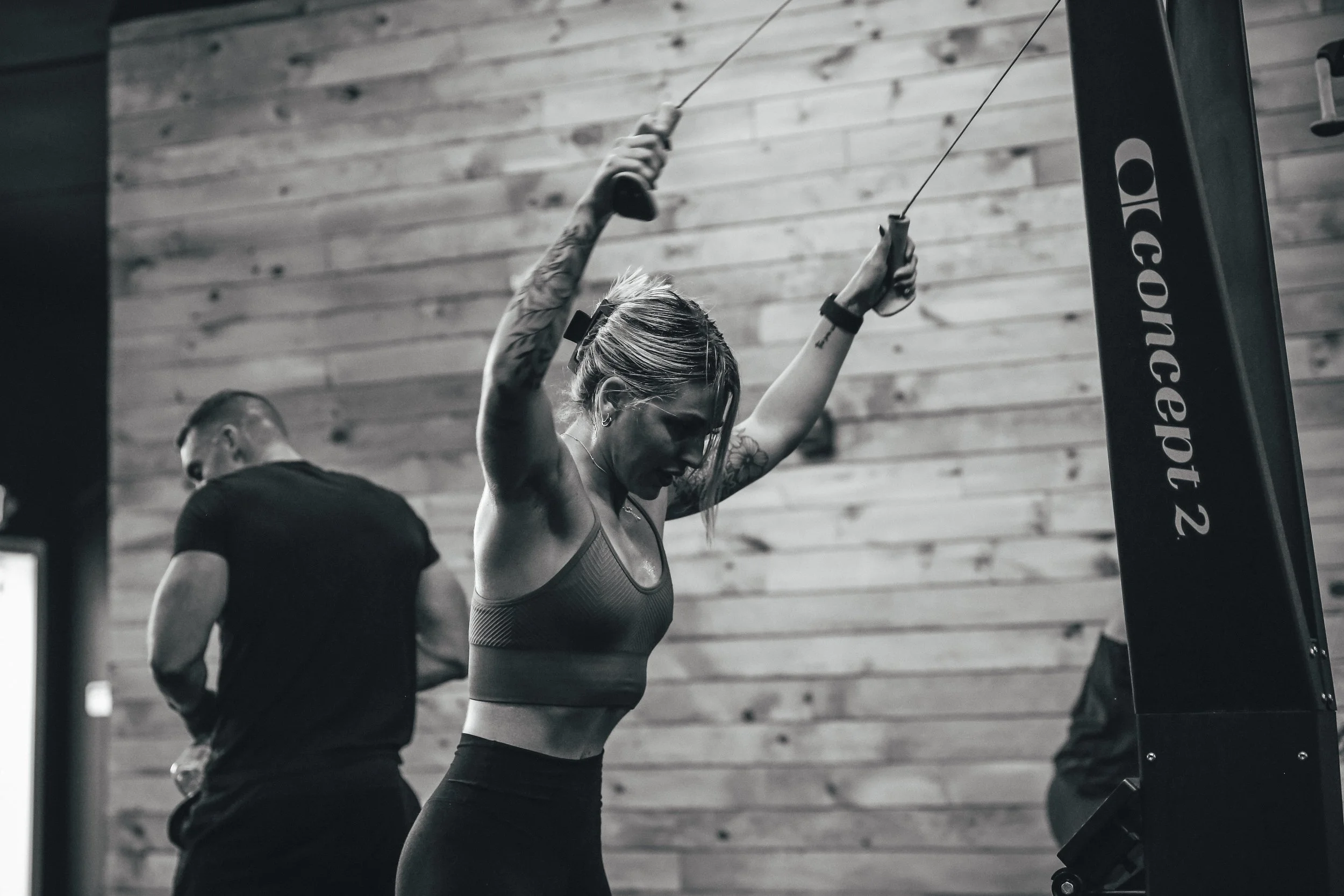 A woman working out on a cable machine in a gym, with a man in the background, both in a black-and-white photograph.