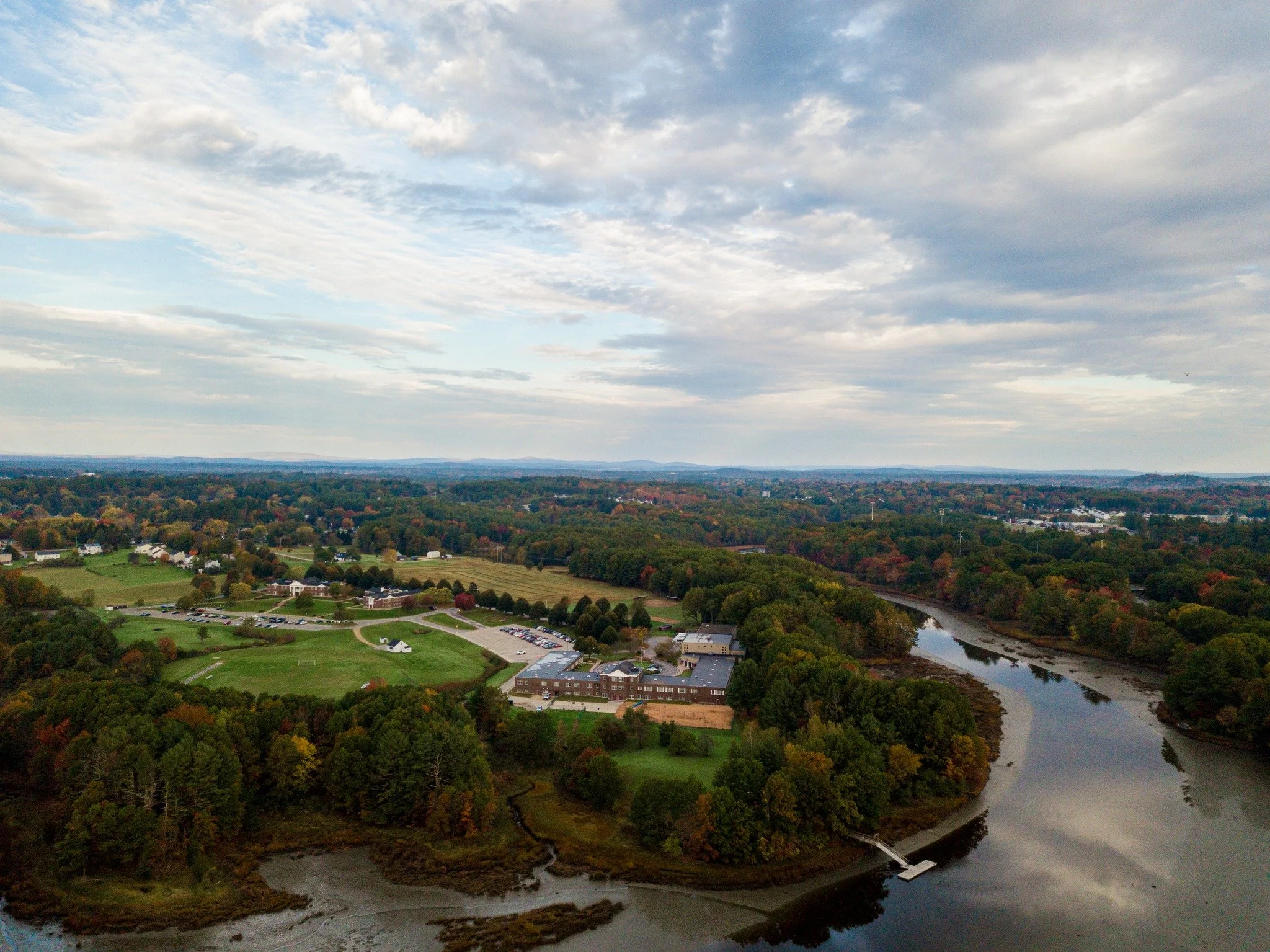 Aerial view of a lush green landscape with a river winding through it, surrounded by trees, fields, and a few buildings under a partly cloudy sky.