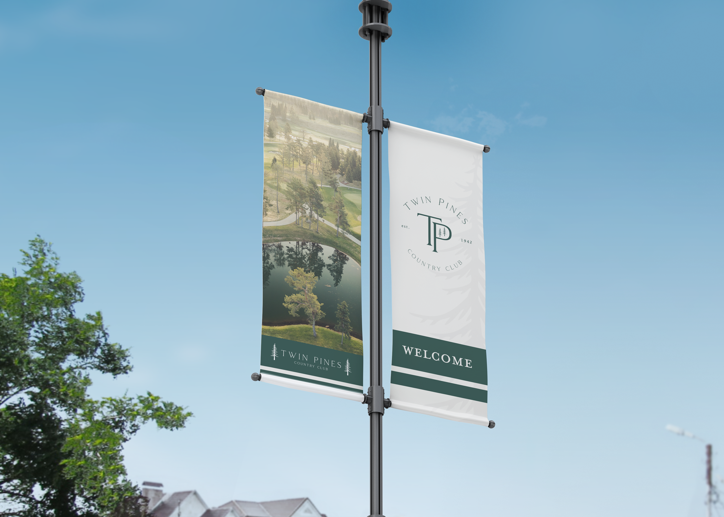 Two banners hanging from a lamppost with the Twin Pines Country Club logo, an aerial view of a golf course and pond, clear blue sky, some trees, and houses in the background.