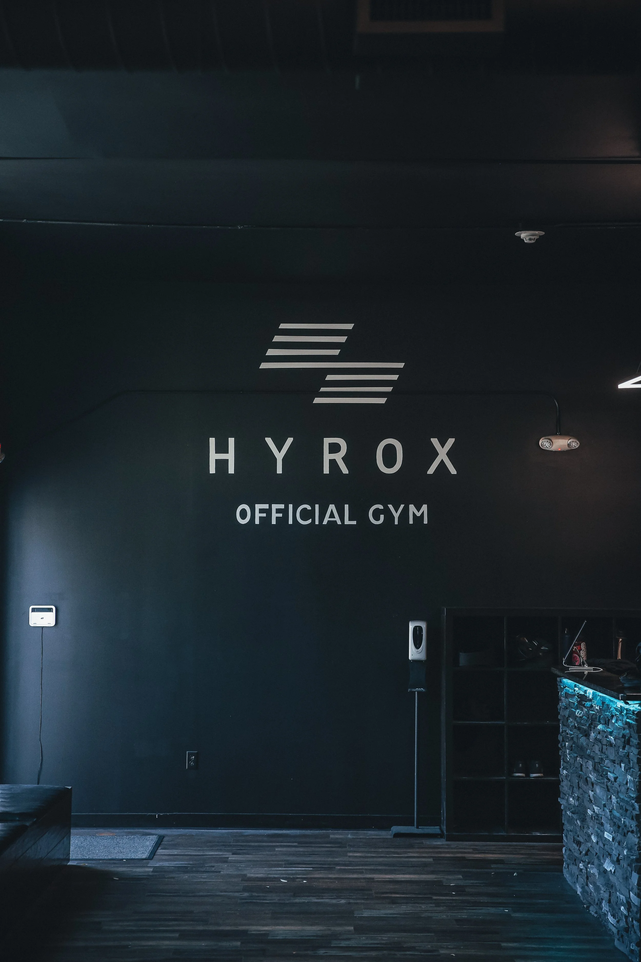 Interior of a gym with a black wall displaying the logo 'HYROX OFFICIAL GYM' and minimalist decor.