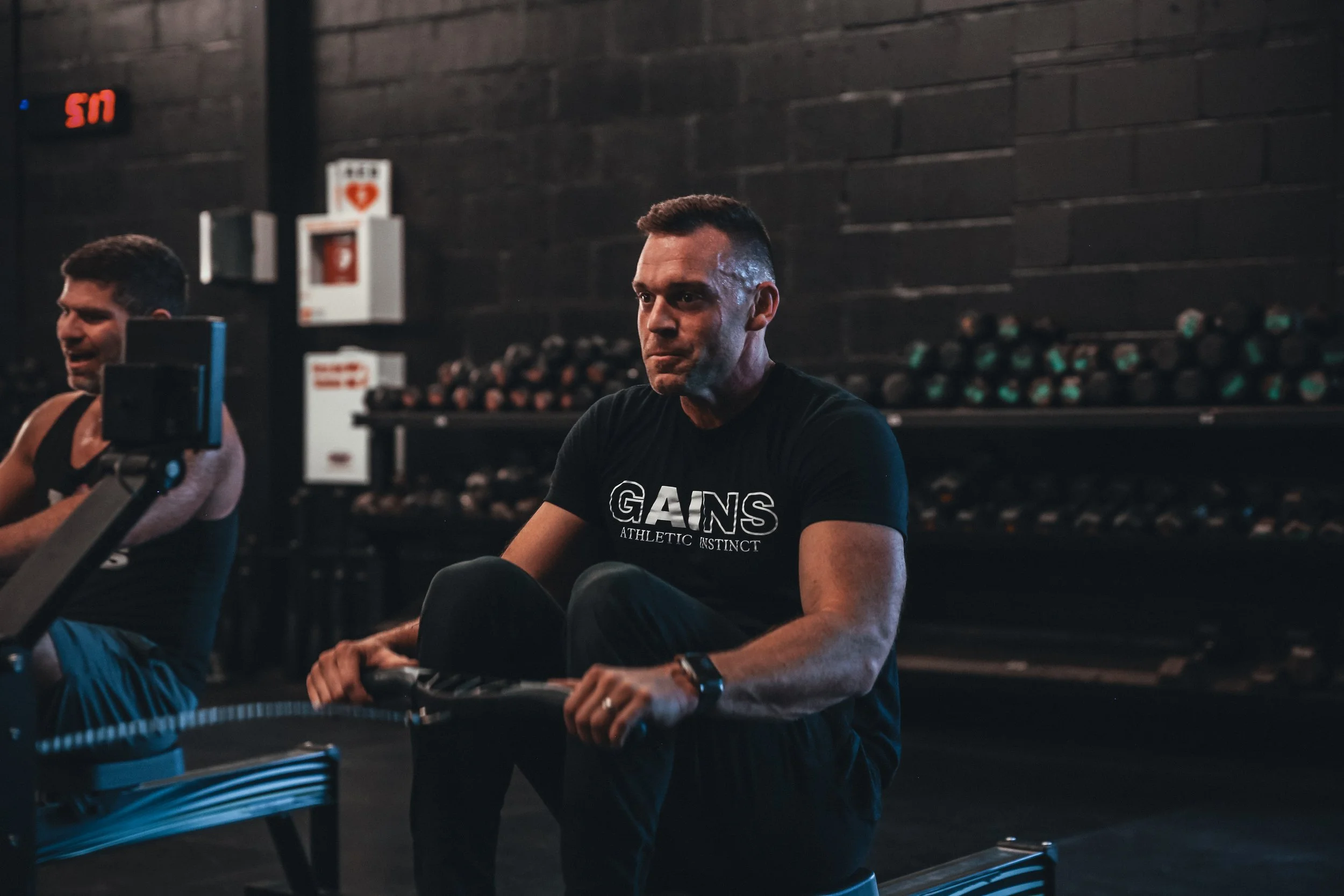 Two men working out on rowing machines in a gym, with black walls and gym equipment in the background.
