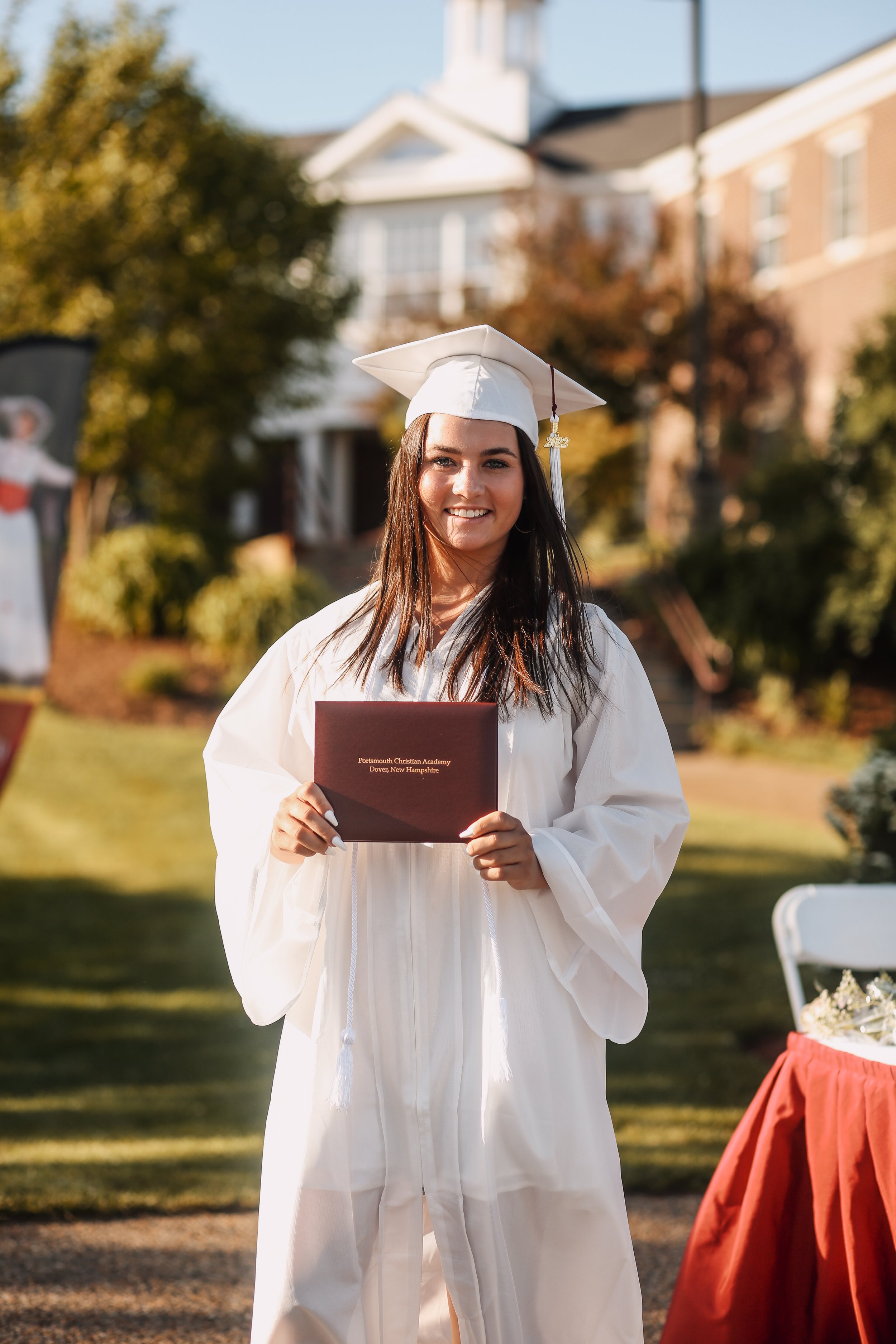 A young woman in a white graduation gown and cap holding a diploma, smiling at a graduation ceremony outdoors.