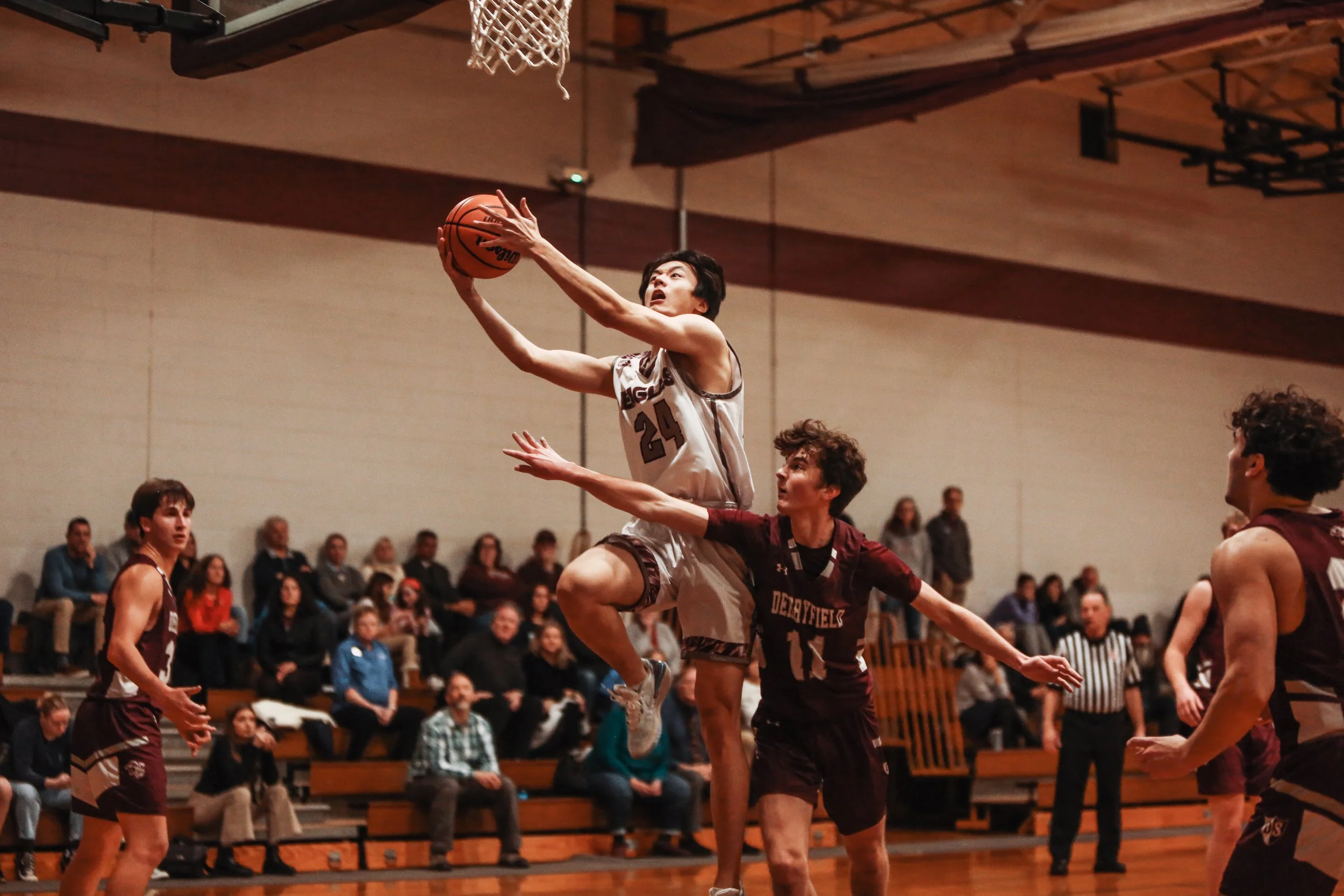A basketball player in a white jersey jumps to shoot the ball while another player in a maroon jersey attempts to block him during a game, with spectators watching in the background.