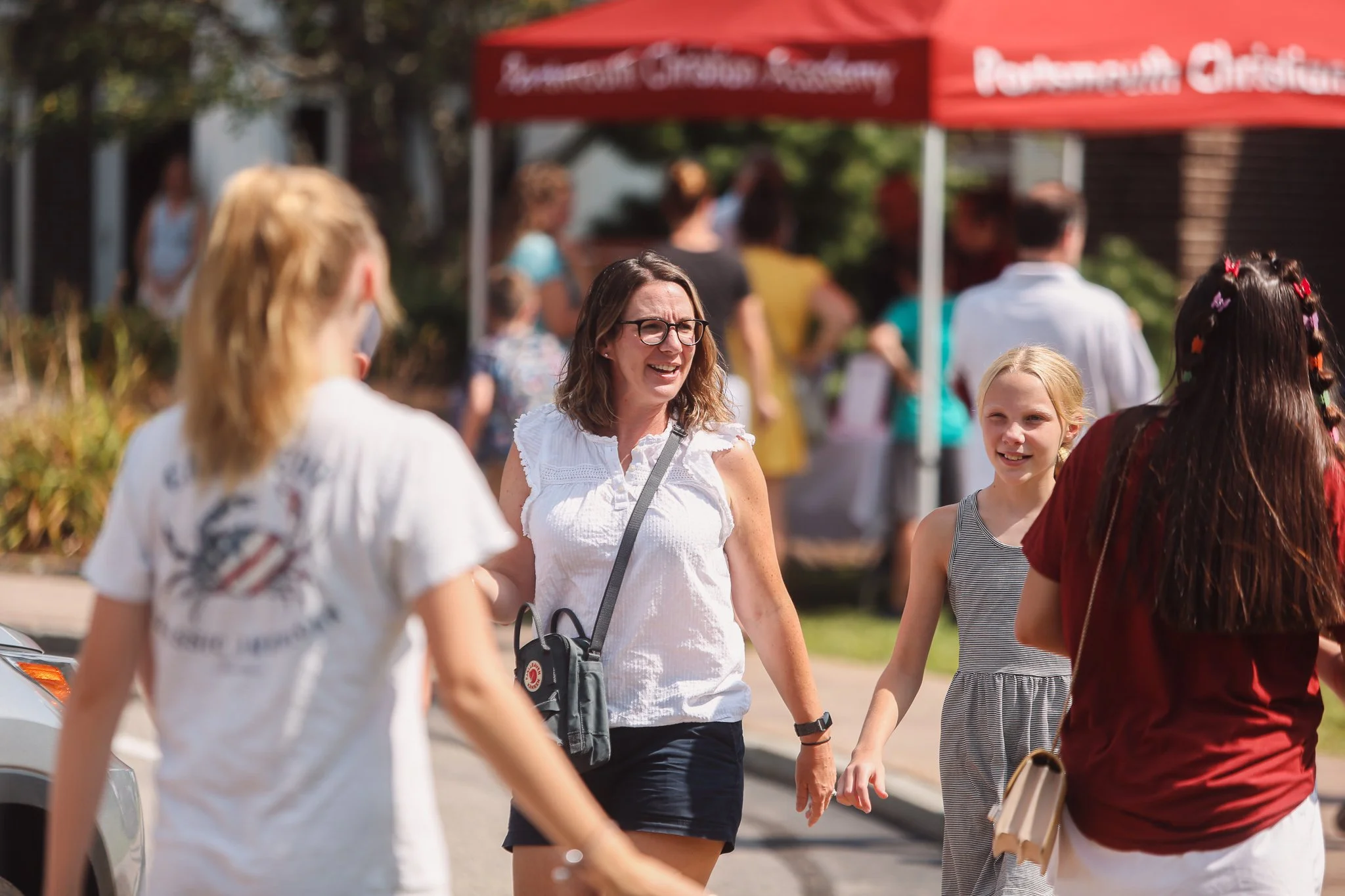 A group of people walking outdoors at an event with a red canopy in the background. The focus is on a woman with glasses, wearing a white top and dark shorts, talking to two young girls.